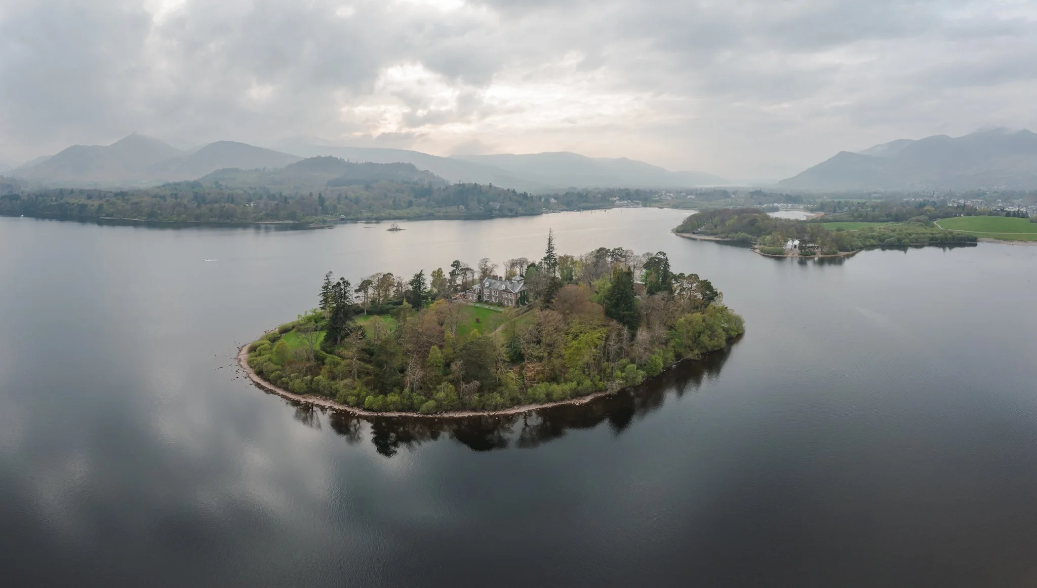 Aerial view of a large lake with an island featuring a mansion, surrounded by lush trees and mountains in the background under cloudy skies.