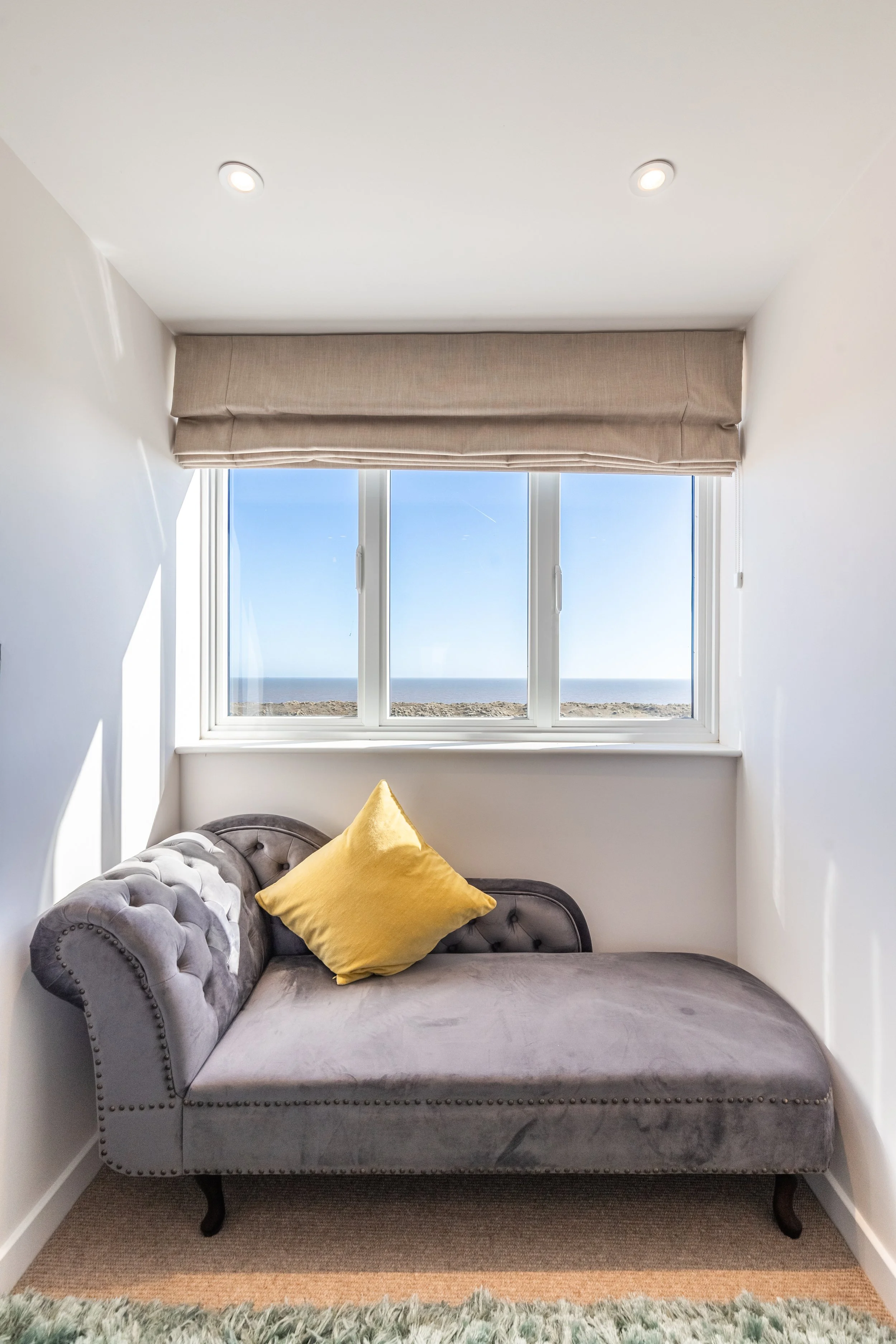 A gray velvet tufted loveseat with black legs and nailhead trim, with a yellow pillow on it. Behind it, a window with a beige roller shade shows a view of the ocean and blue sky. The room has white walls and a beige carpet, with a gray and cream shag