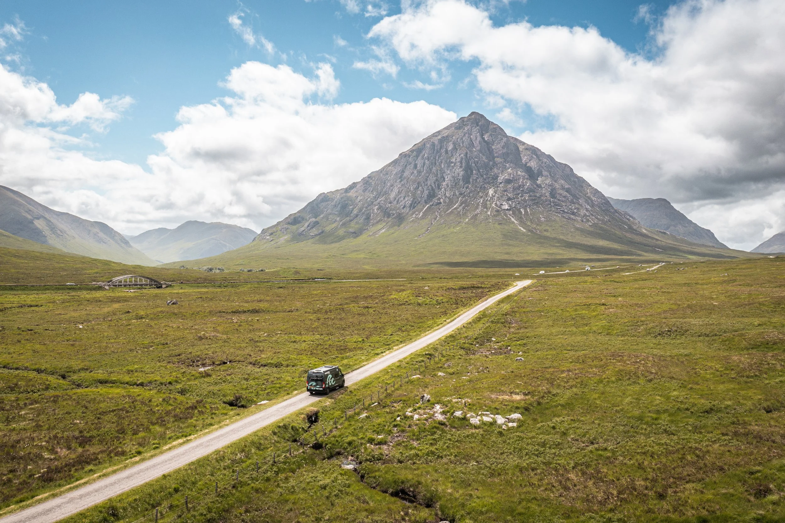 A bus traveling along a winding road through a green landscape with a large, rugged mountain in the background and partly cloudy sky.
