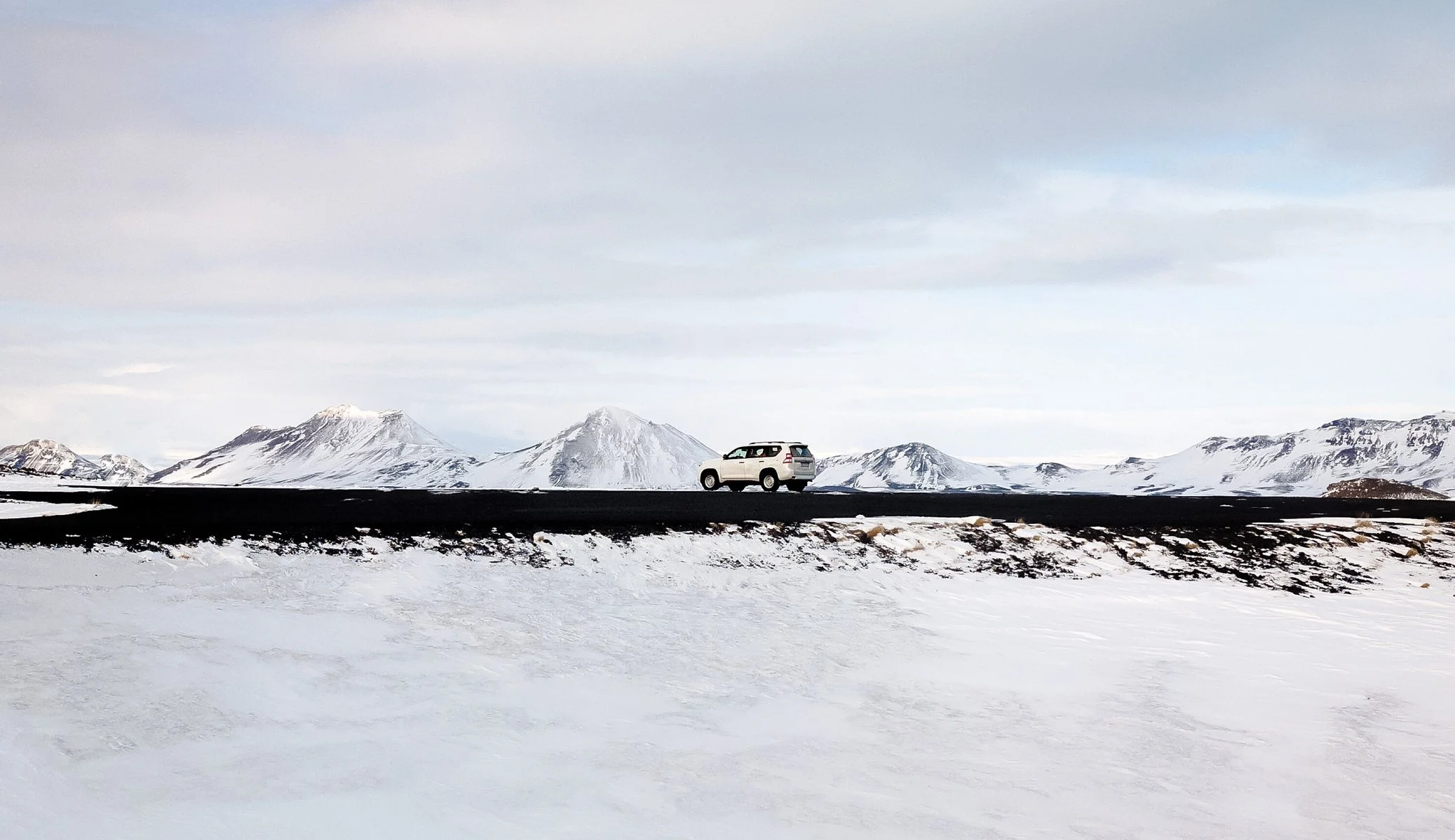 A white SUV driving on a black asphalt road through a snowy landscape with snow-capped mountains under a cloudy sky.