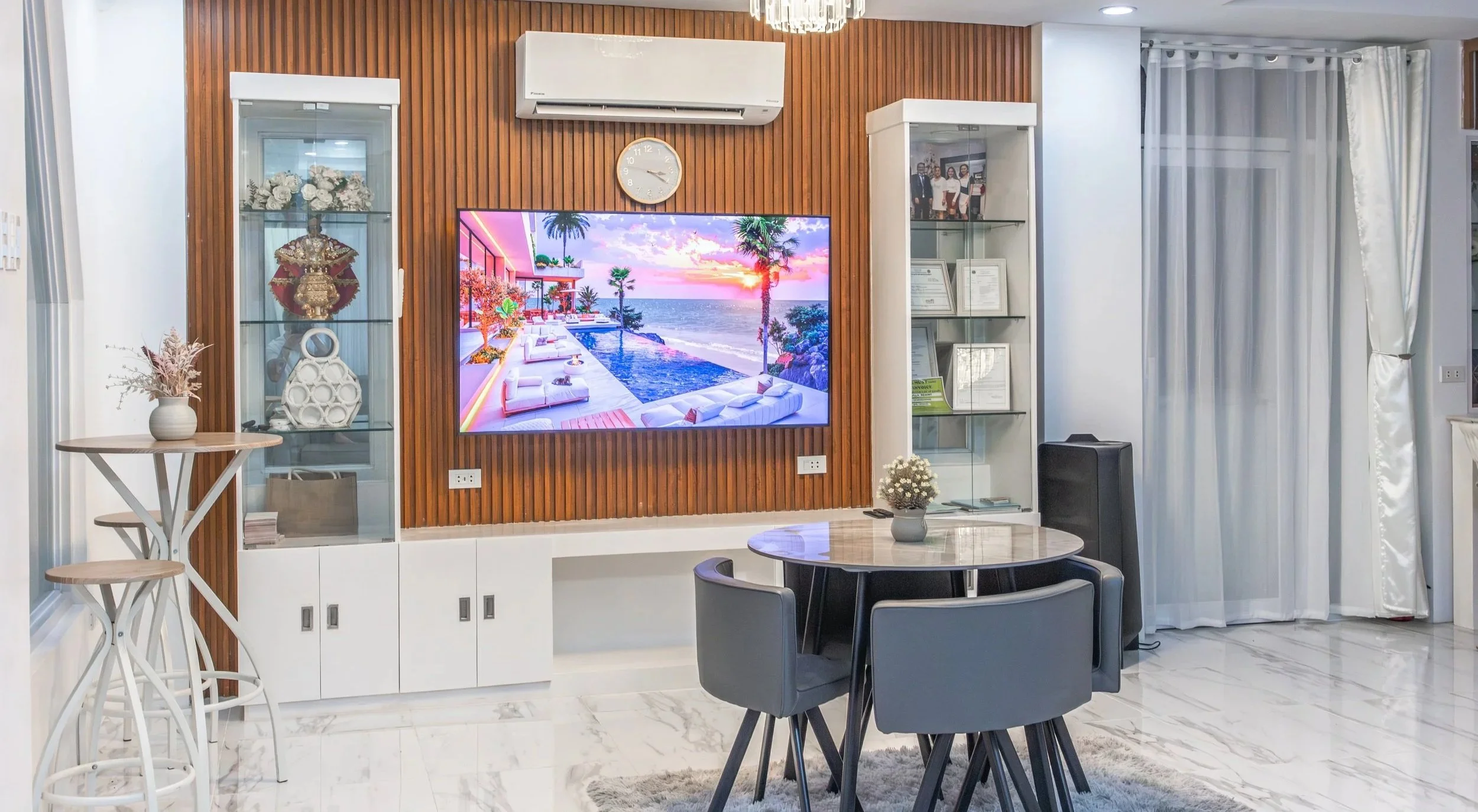 Living room with a wooden accent wall, mounted TV displaying a tropical resort scene, white air conditioner, glass display cabinets with decorative items, round table with a vase and flowers, and dining chairs, with white curtains on a window to the right.