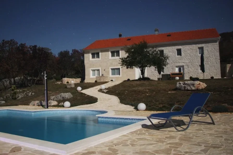 Nighttime view of a house with a swimming pool in the foreground, a blue lounge chair, a walking path with white spherical lights, and a starry sky.