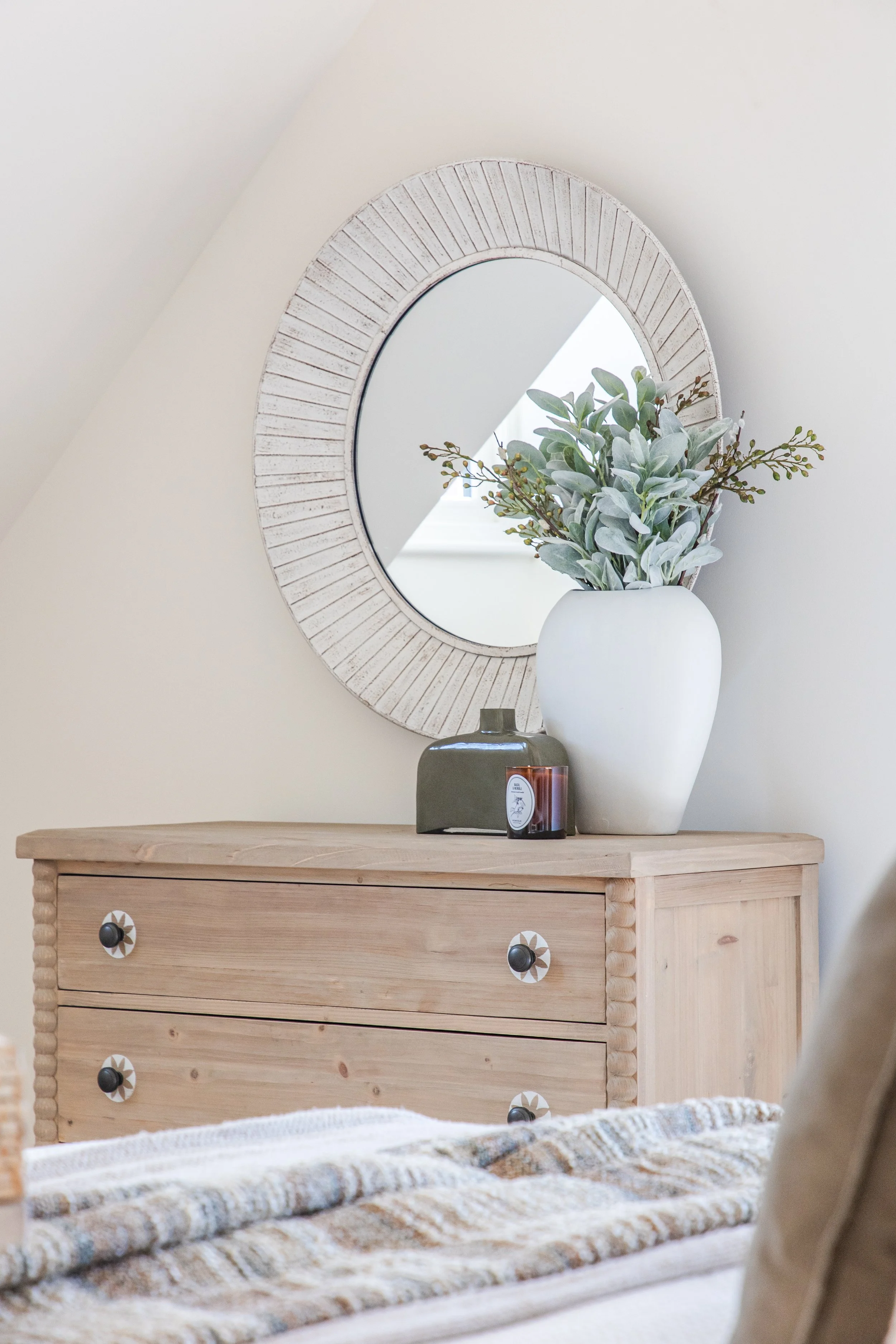 A wooden dresser with three drawers, featuring decorative black knobs, is placed against a white wall. On top of the dresser, there is a white vase with green foliage, a small greenish object, and a brown candle. A large, round mirror with a textured