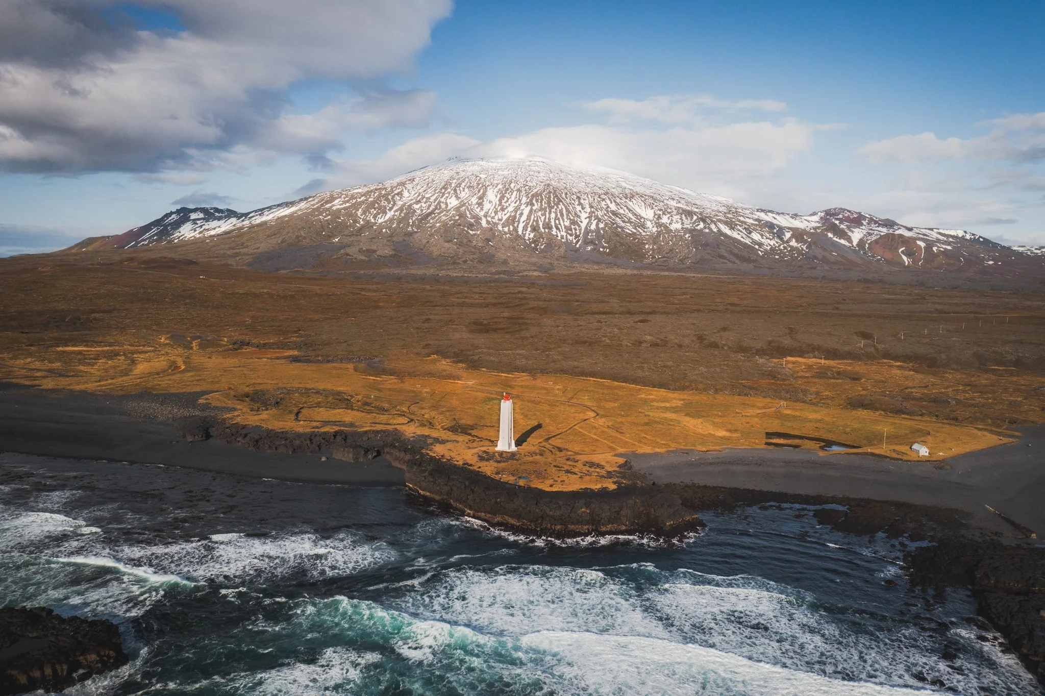 A snow-capped volcano with barren brown terrain in front of a cloudy sky, near the ocean with dark rocky shores and waves.