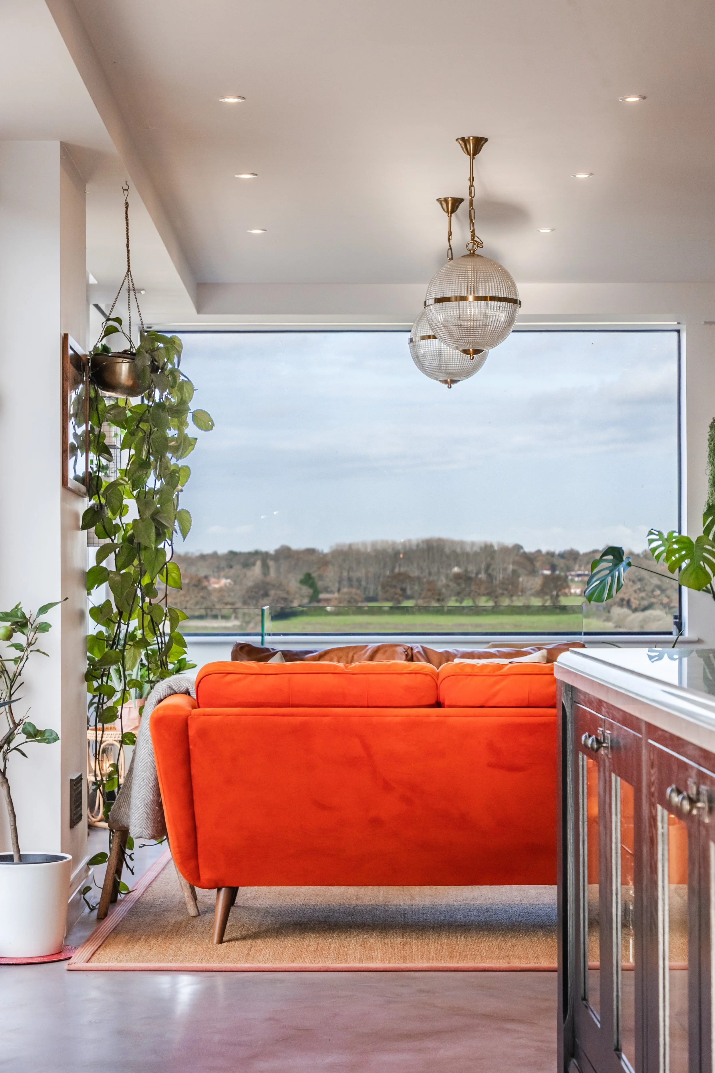 Living room with a bright orange couch, a potted plant on the left, a built-in cabinet on the right, a large window showing a landscape of trees and hills, and a hanging light fixture with three globes.