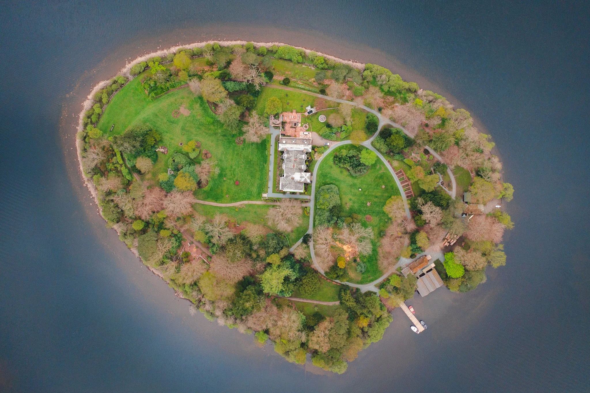 Aerial view of a small green island with a large house, trees, pathways, and a lawn surrounded by calm water, with boats docked at a pier.