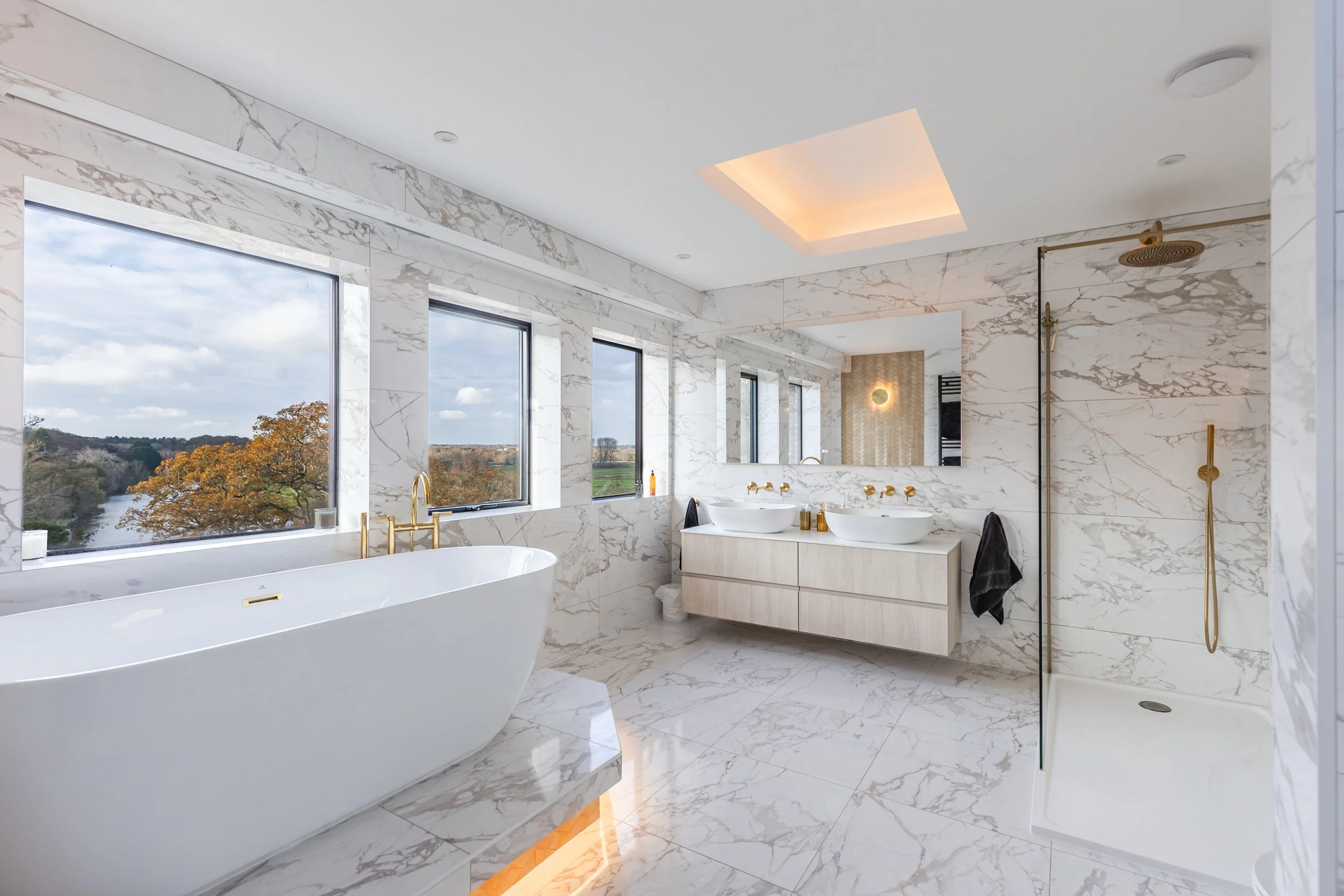 Luxurious bathroom with white marble walls and floors, a freestanding bathtub, double sinks with gold fixtures, large windows showing an outdoor landscape, a mirror, and a shower area with a gold showerhead.