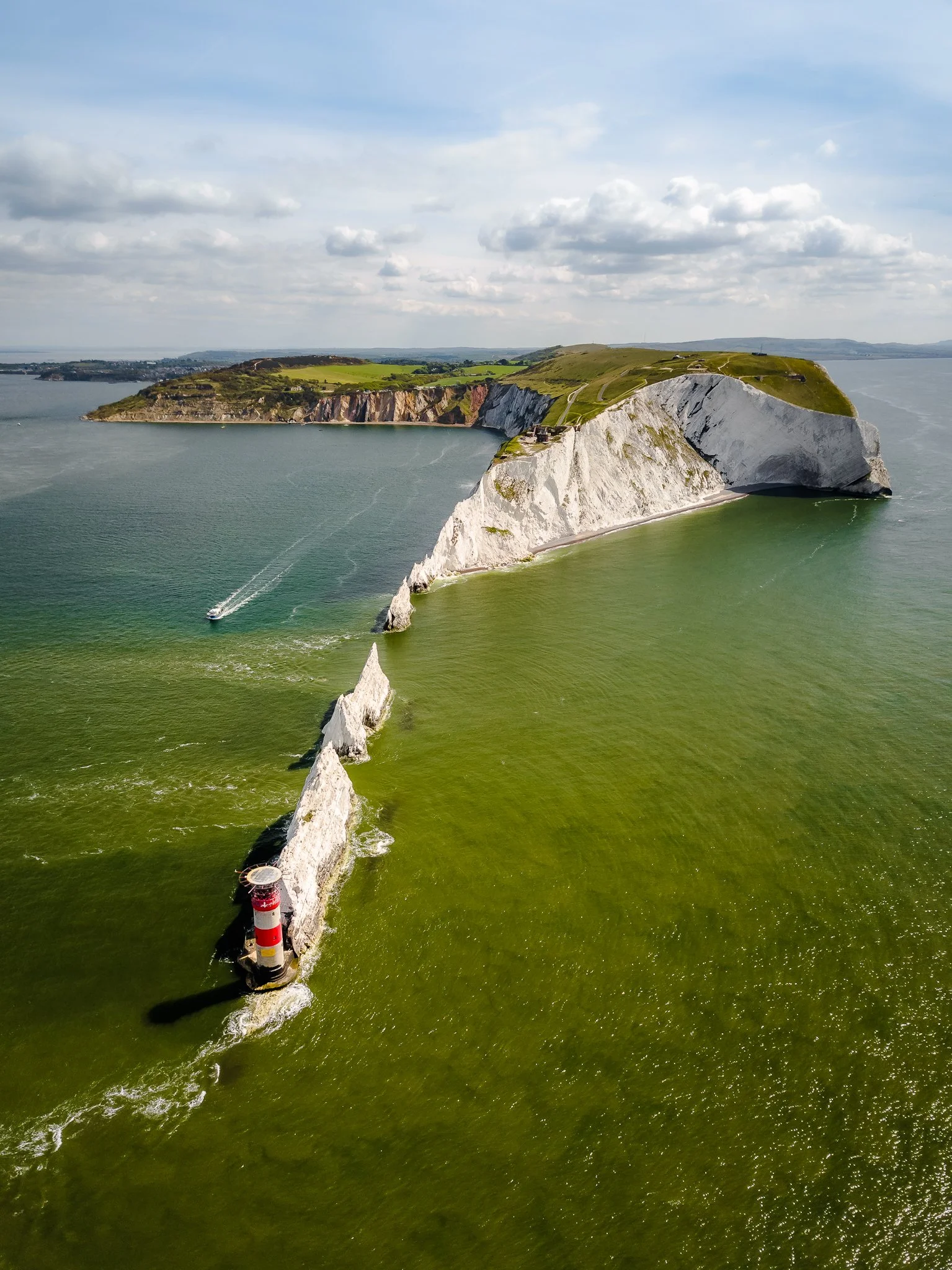 Aerial view of a white chalk cliff protruding into the ocean, with a red and white lighthouse at its base, and a boat sailing nearby. The scene shows a partly cloudy sky and green waters.