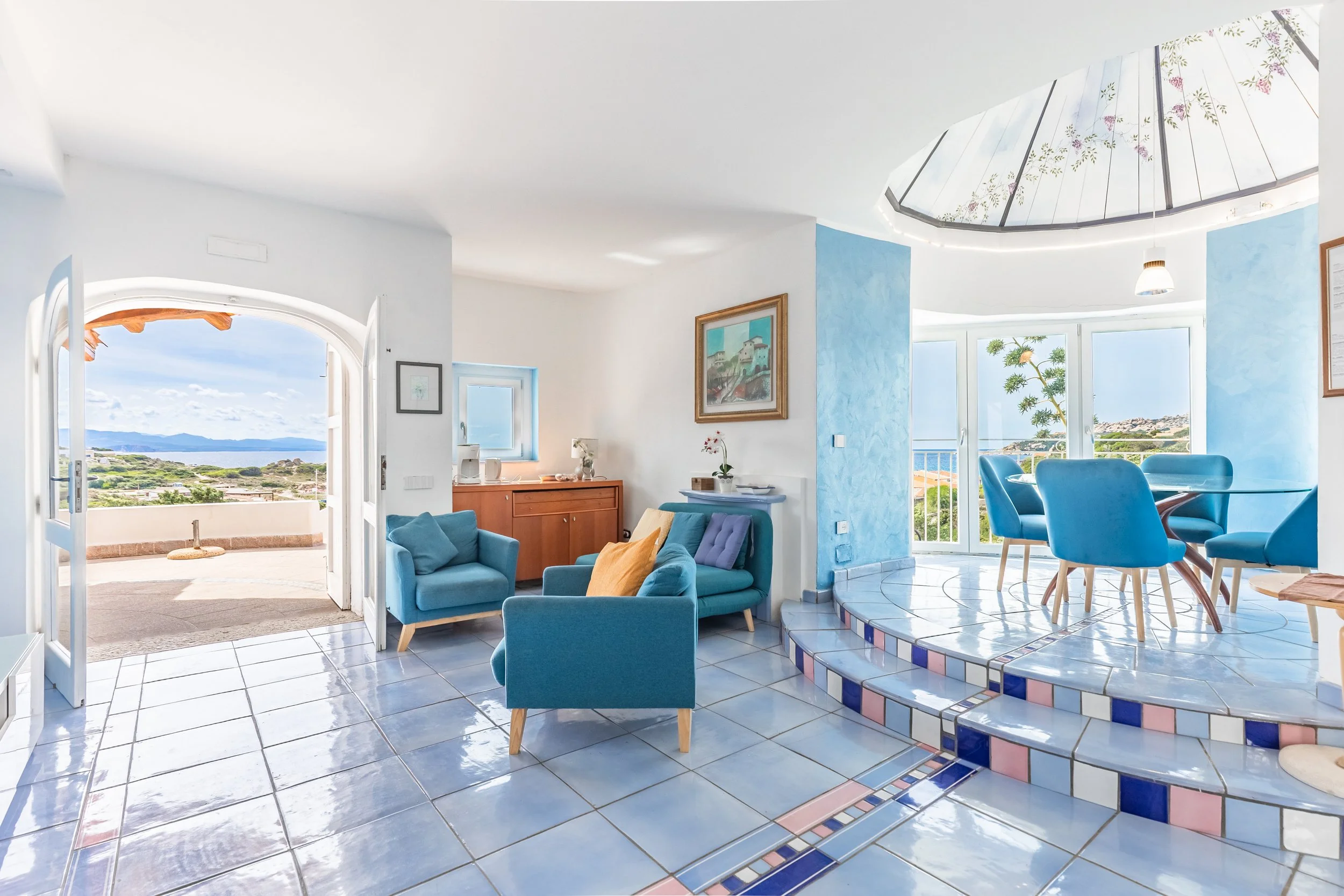 Bright living room with blue furniture, artwork, and a view of a scenic landscape through multiple windows and a glass ceiling.