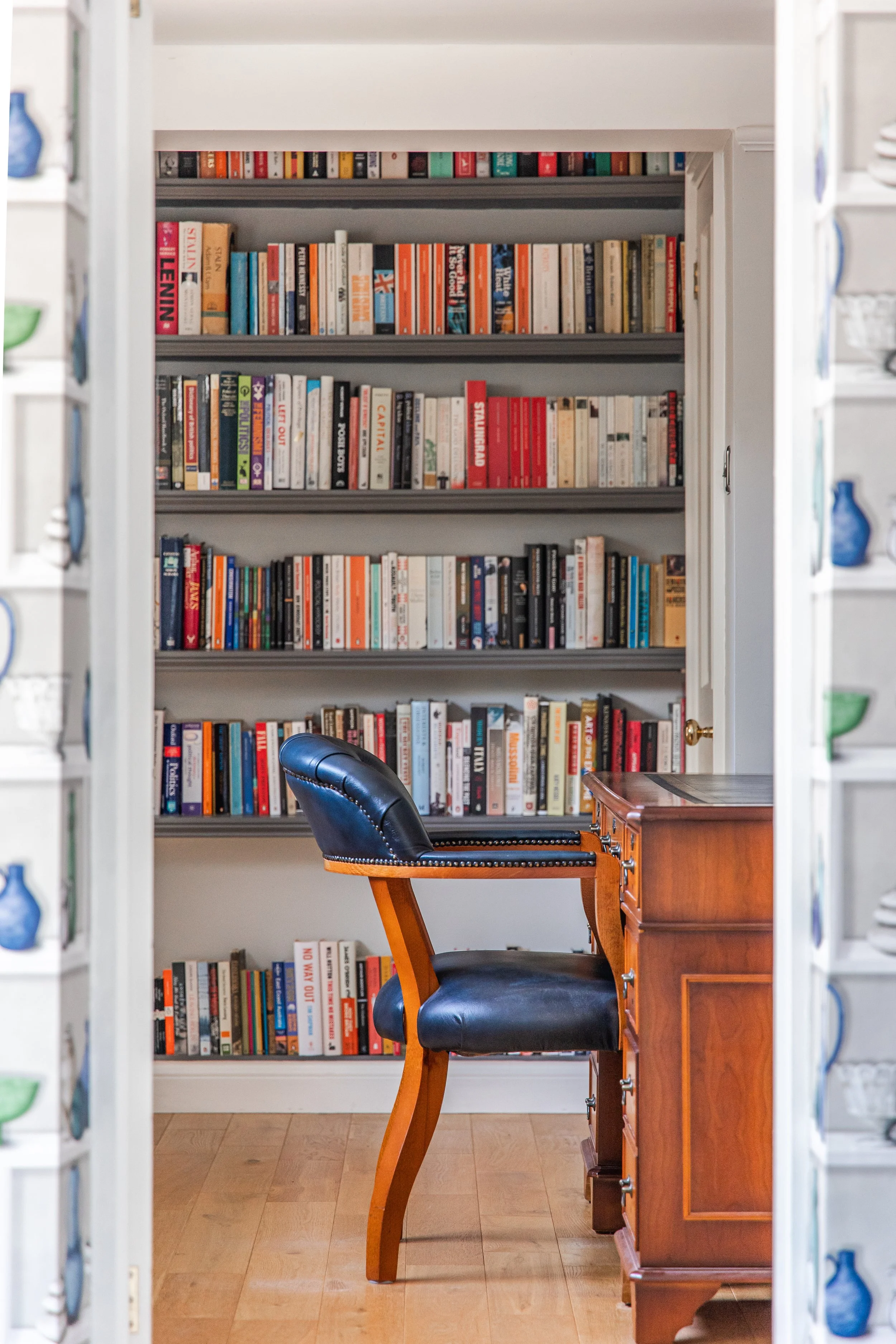 View of a living room or study with a large bookshelf filled with books, a wooden desk, and a black leather office chair.