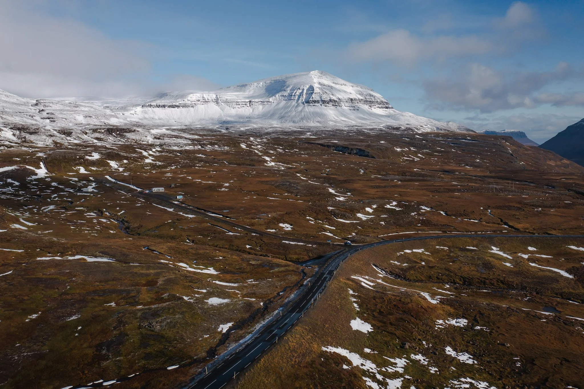 A mountain covered in snow with a winding road in the foreground and patches of snow on brown terrain under a partly cloudy sky.