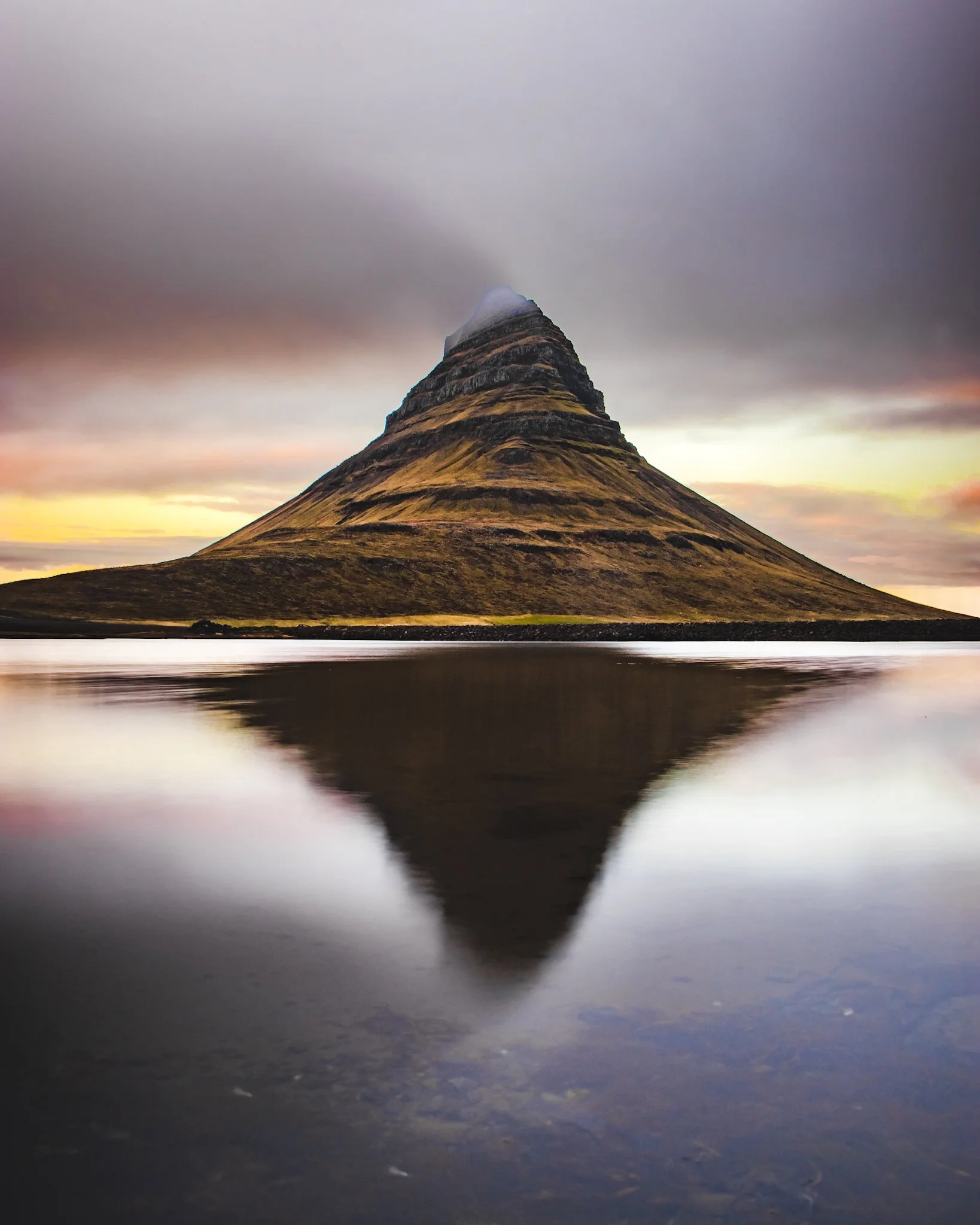 A mountain with a pointed peak partially covered by clouds, reflected in a body of water at sunset with a cloudy sky in the background.