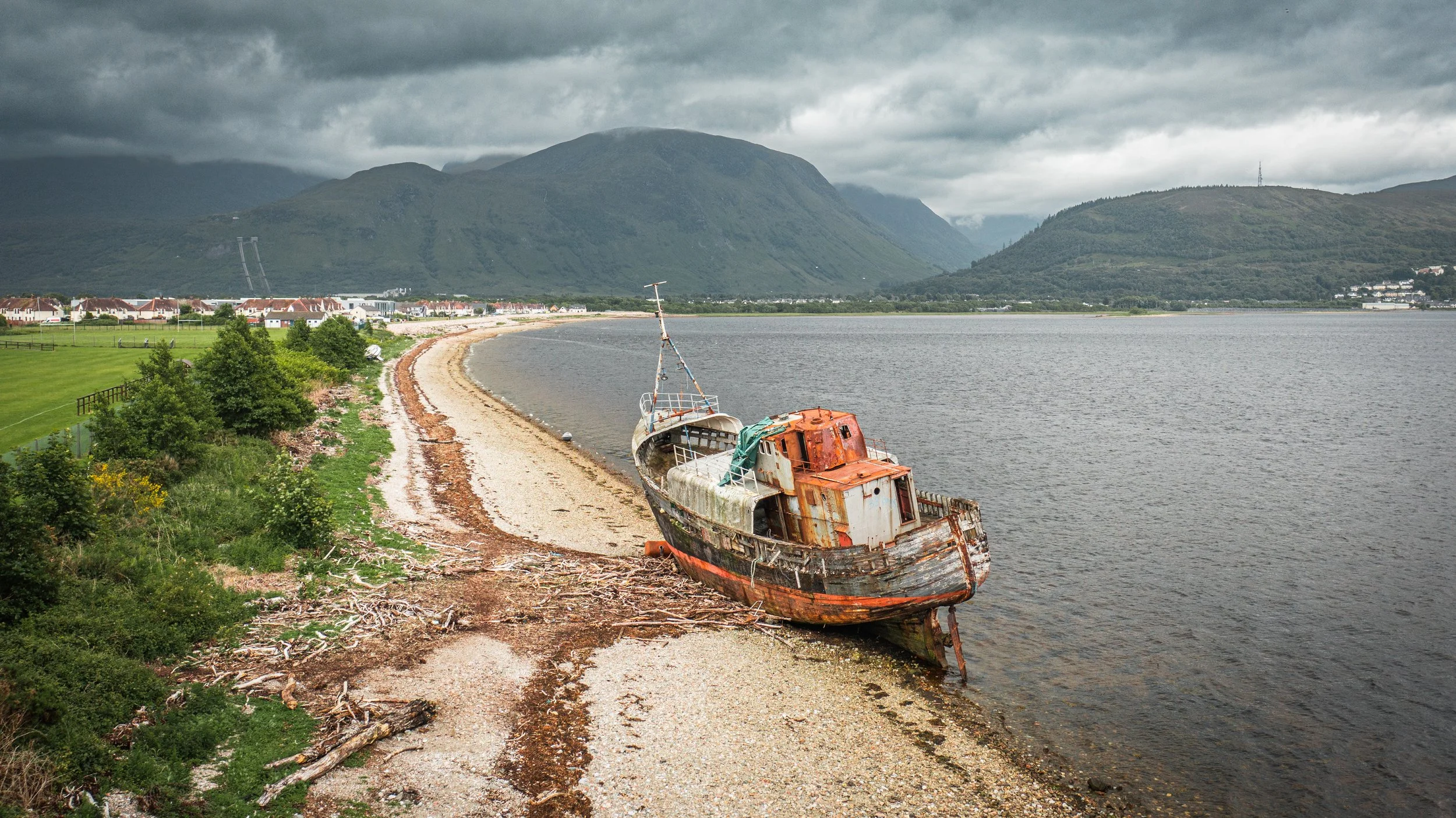 An abandoned boat with rusty and damaged hull on a pebbled shoreline, with a small town and green hills in the background under a cloudy sky.