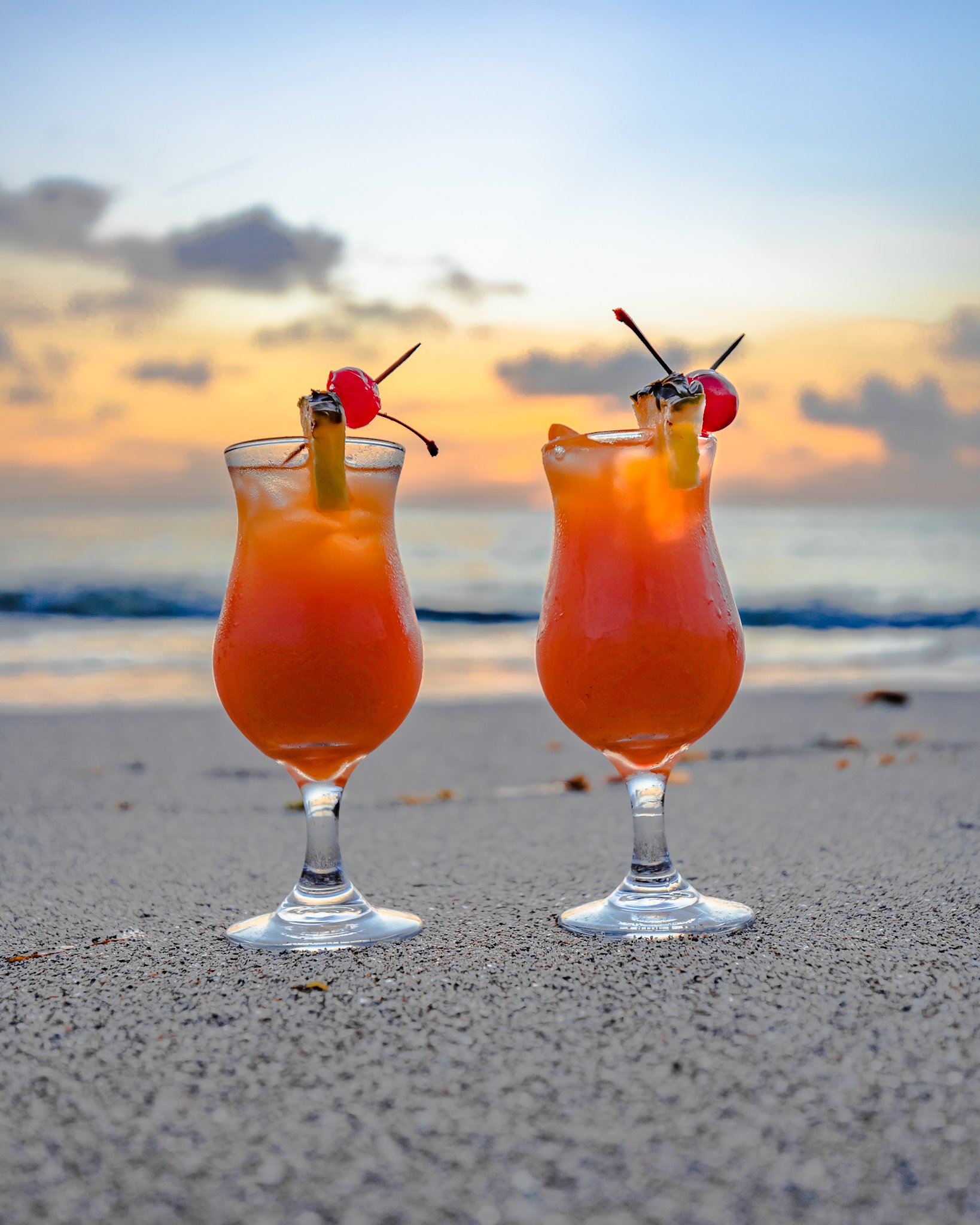 Two tropical cocktails with cherries and pineapple slices on a sandy beach at sunset.