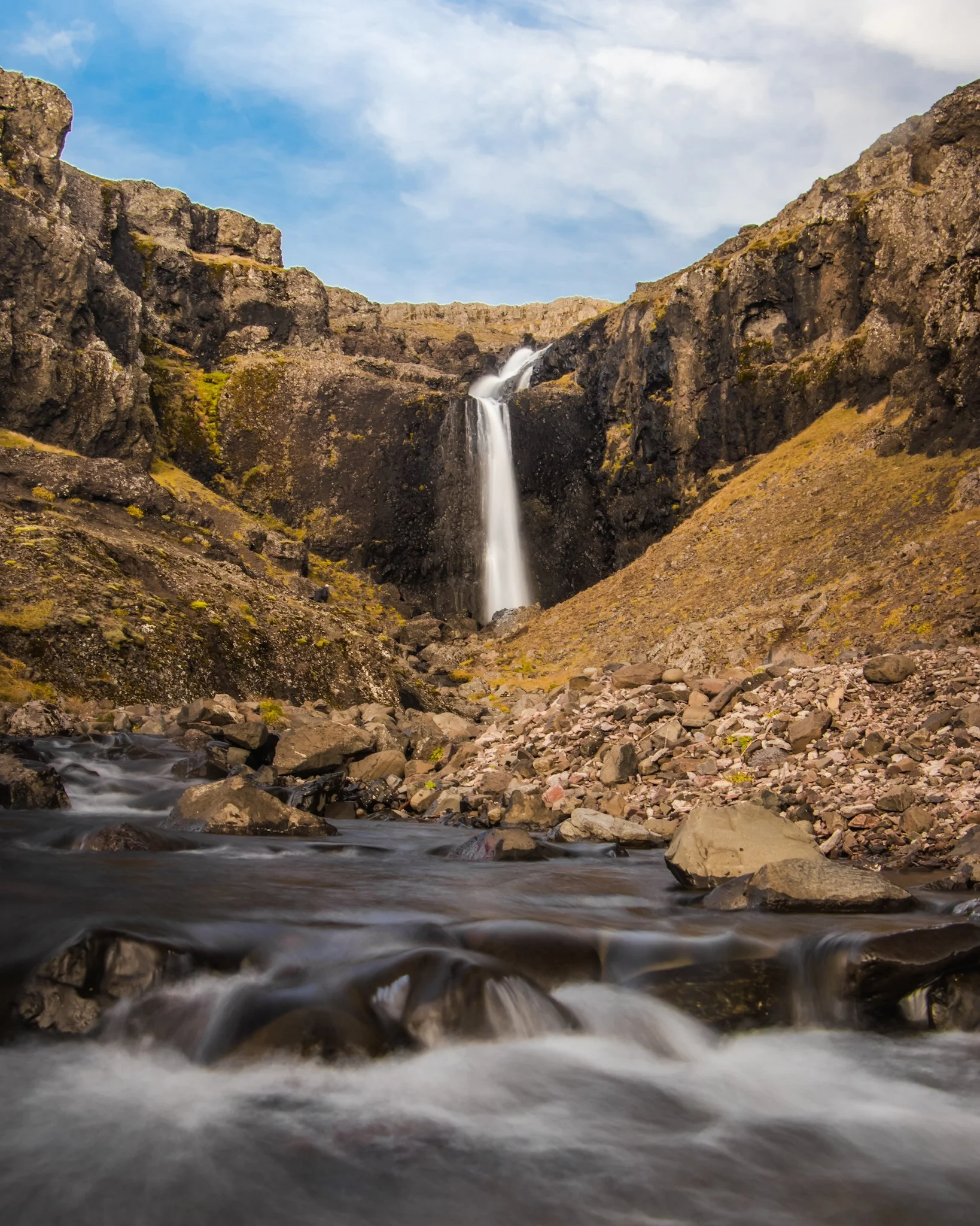 A waterfall flowing down a rocky mountain into a stream with rocks and pebbles.