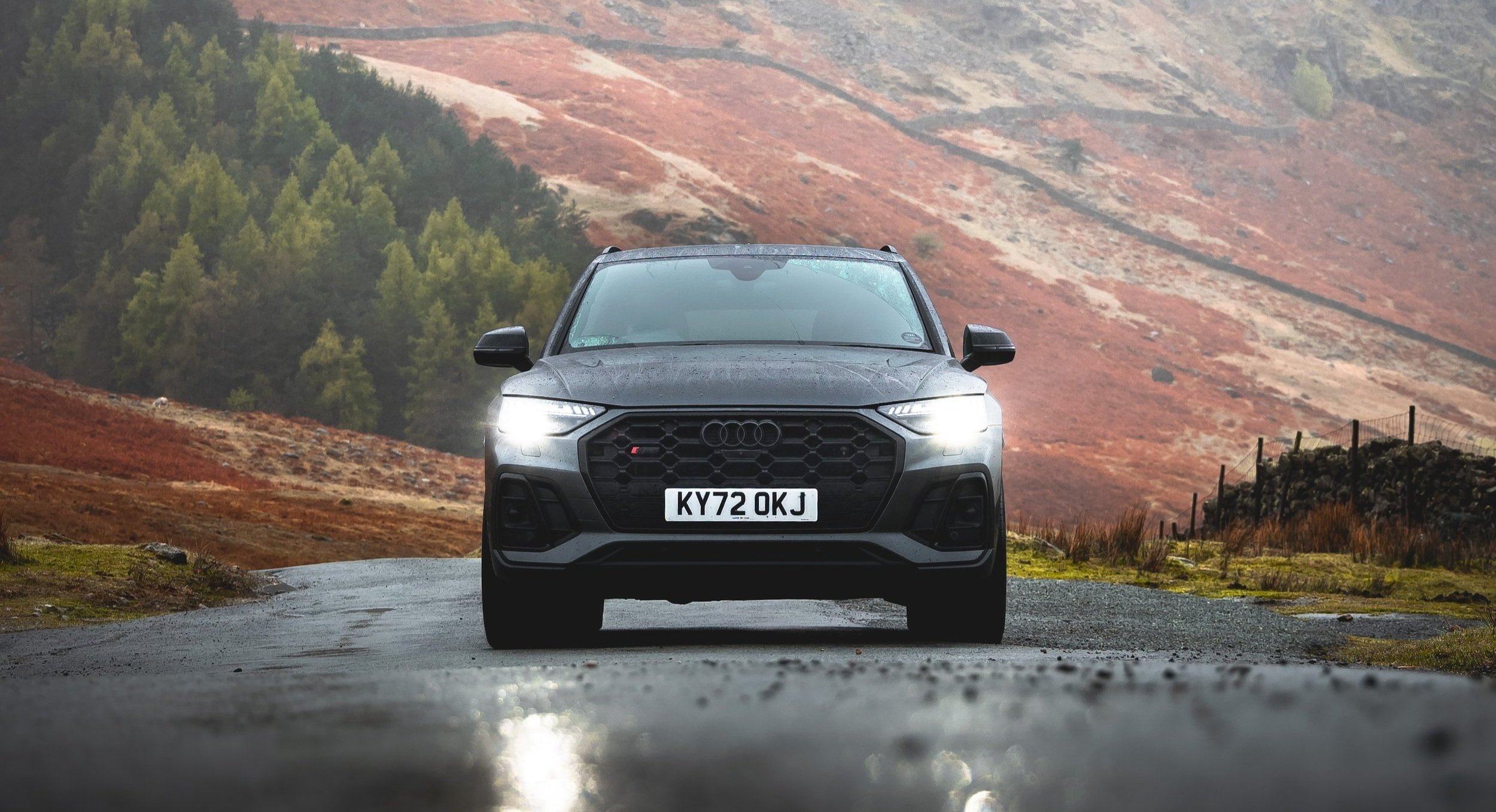 A black Audi SUV driving on a wet road with mountains and green trees in the background.