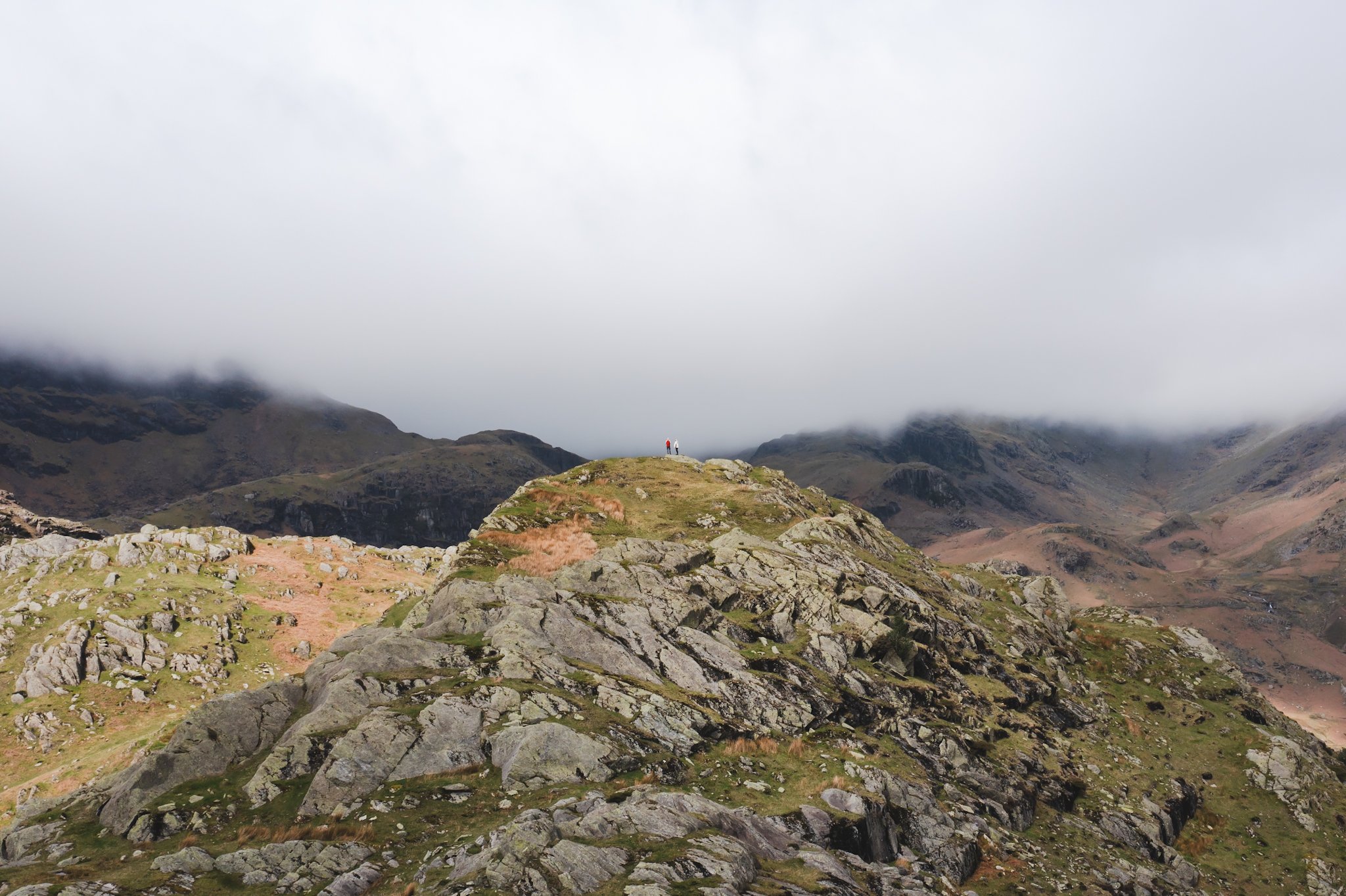 Mountain landscape with rocky terrain and a person in red on a small hilltop, surrounded by misty mountains under cloudy sky.
