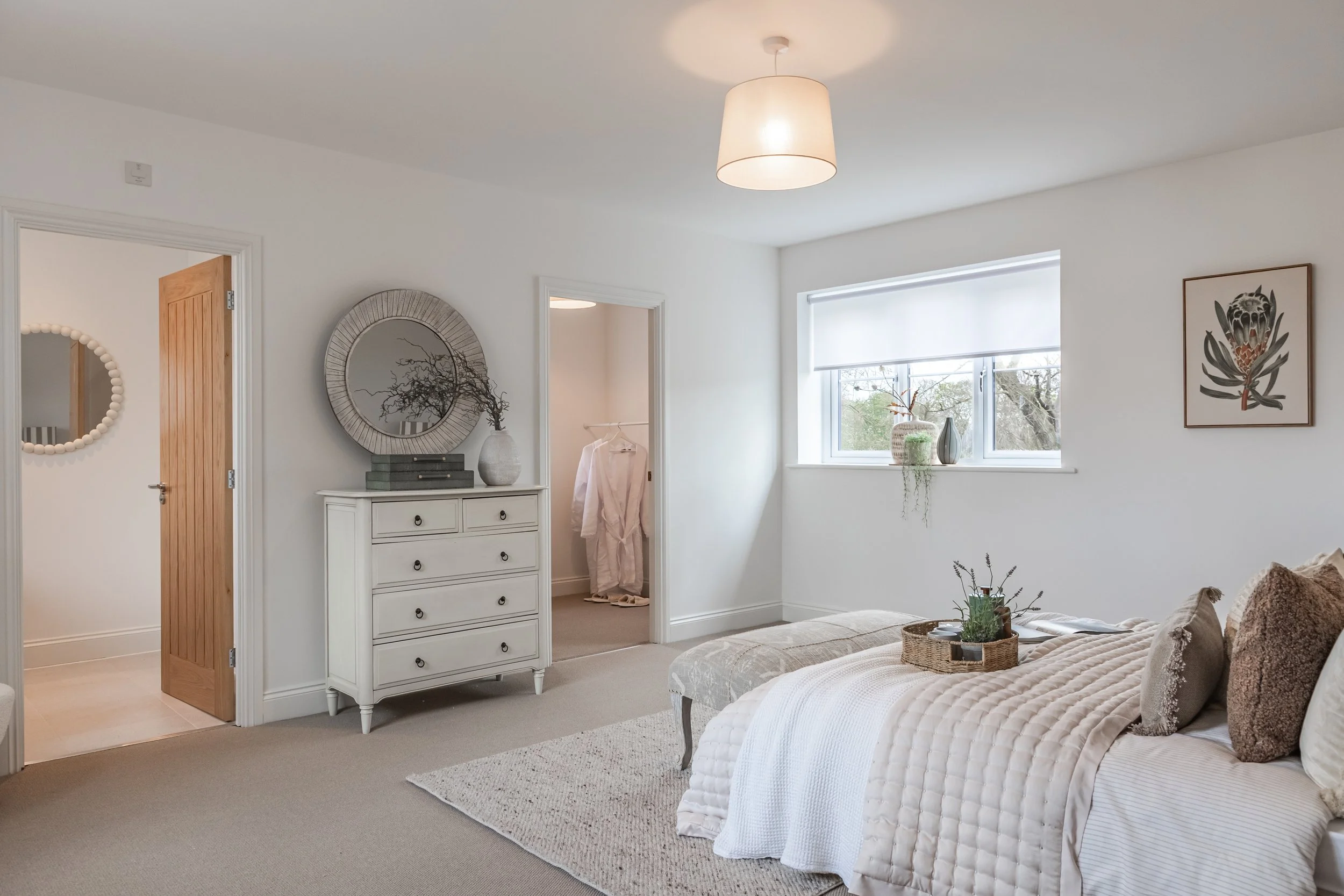A cozy bedroom with a large window, white walls, a bed with beige quilts and pillows, a woven basket, and artwork on the wall, with a dresser and mirror nearby