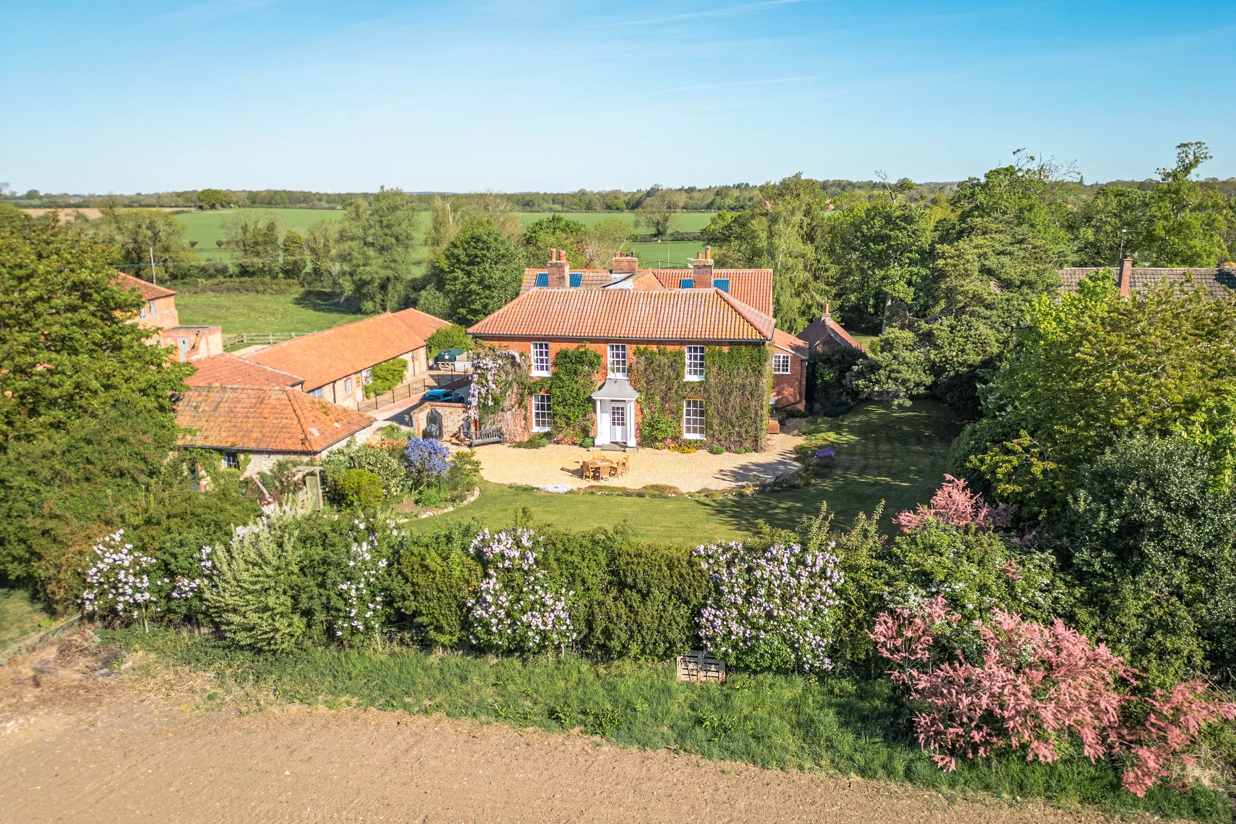 Aerial view of a countryside with red brick houses, green trees, and fields in the background under a blue sky.
