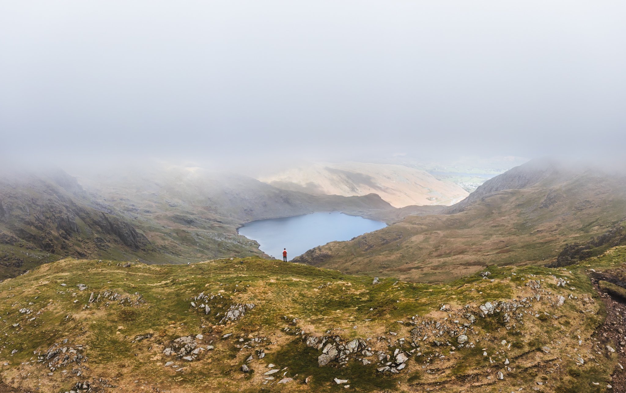 A person standing on a mountain ridge looking at a lake below surrounded by mountains and an overcast sky.