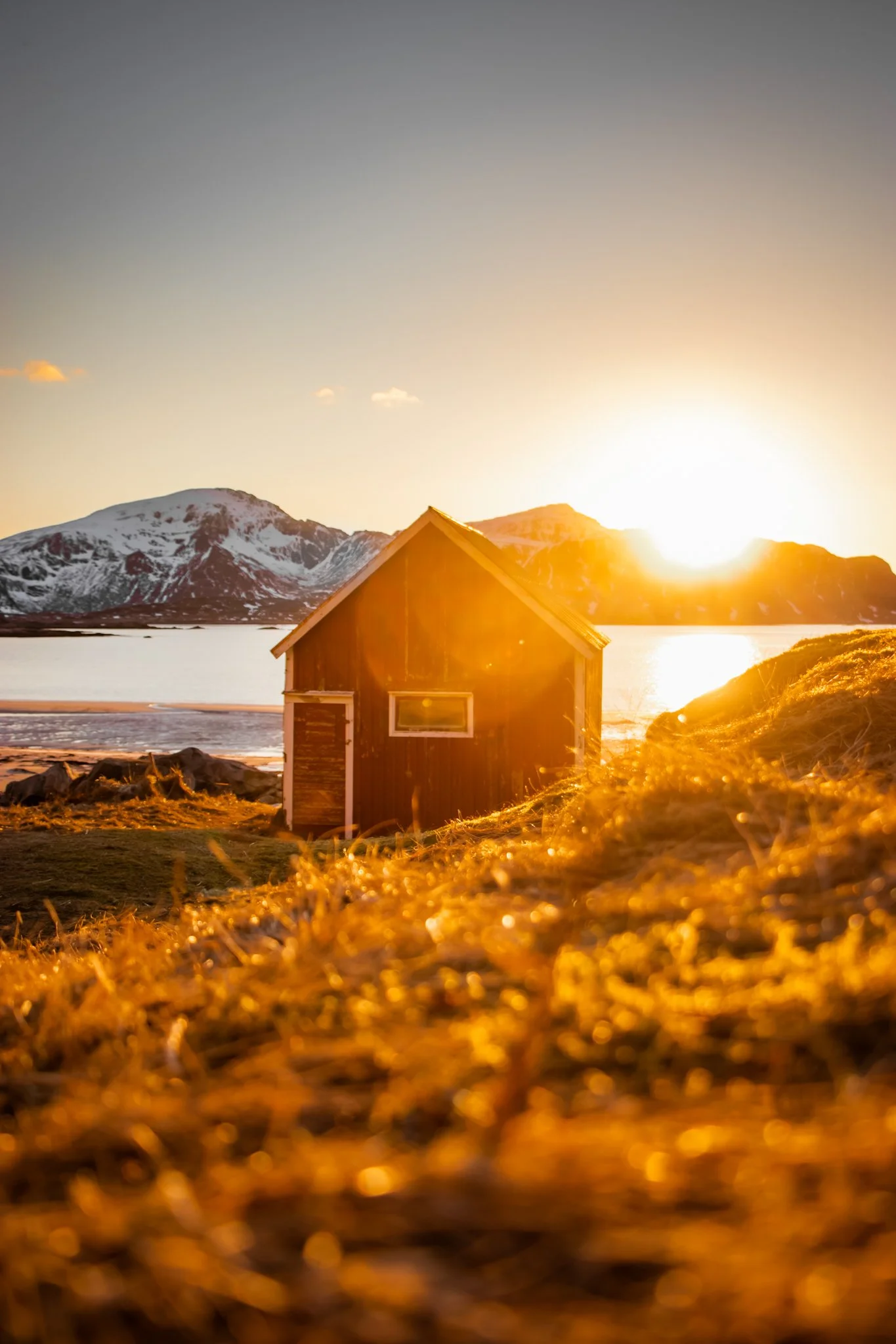 Sunset over snow-capped mountains with a small wooden cabin in the foreground, glowing with warm sunlight.