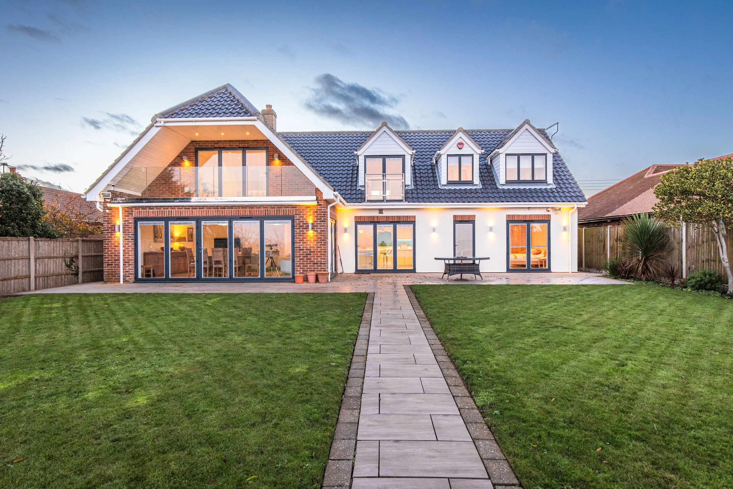 Modern two-story house with a backyard, large sliding glass doors, brick and white exterior, multiple dormer windows, and a paved pathway leading to a patio with a table and chairs, surrounded by a grassy lawn and trees.