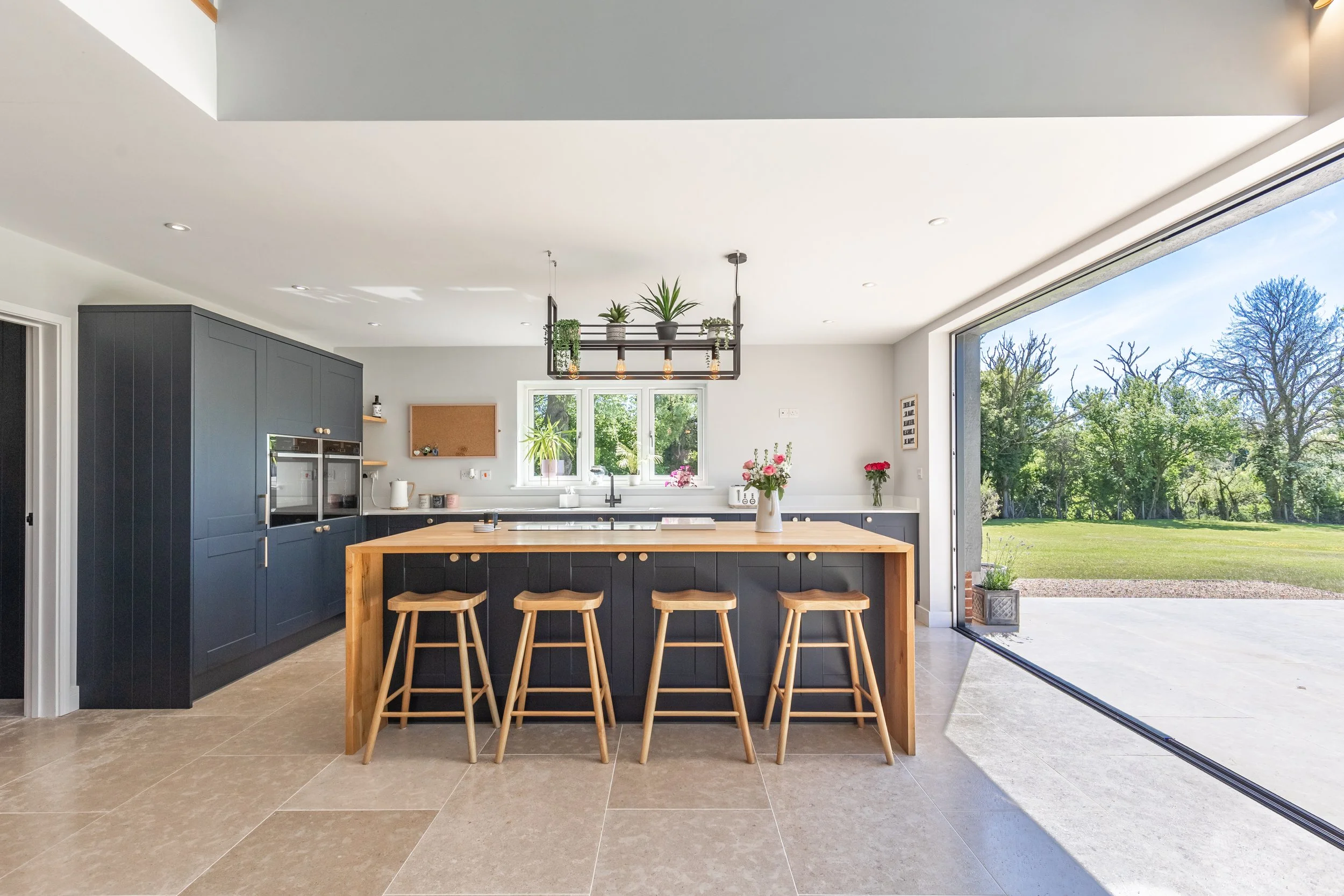 Modern kitchen with dark blue cabinets, a wooden island with bar stools, flower vases, and plants above, large sliding glass door opening to a outdoor green landscape with trees.