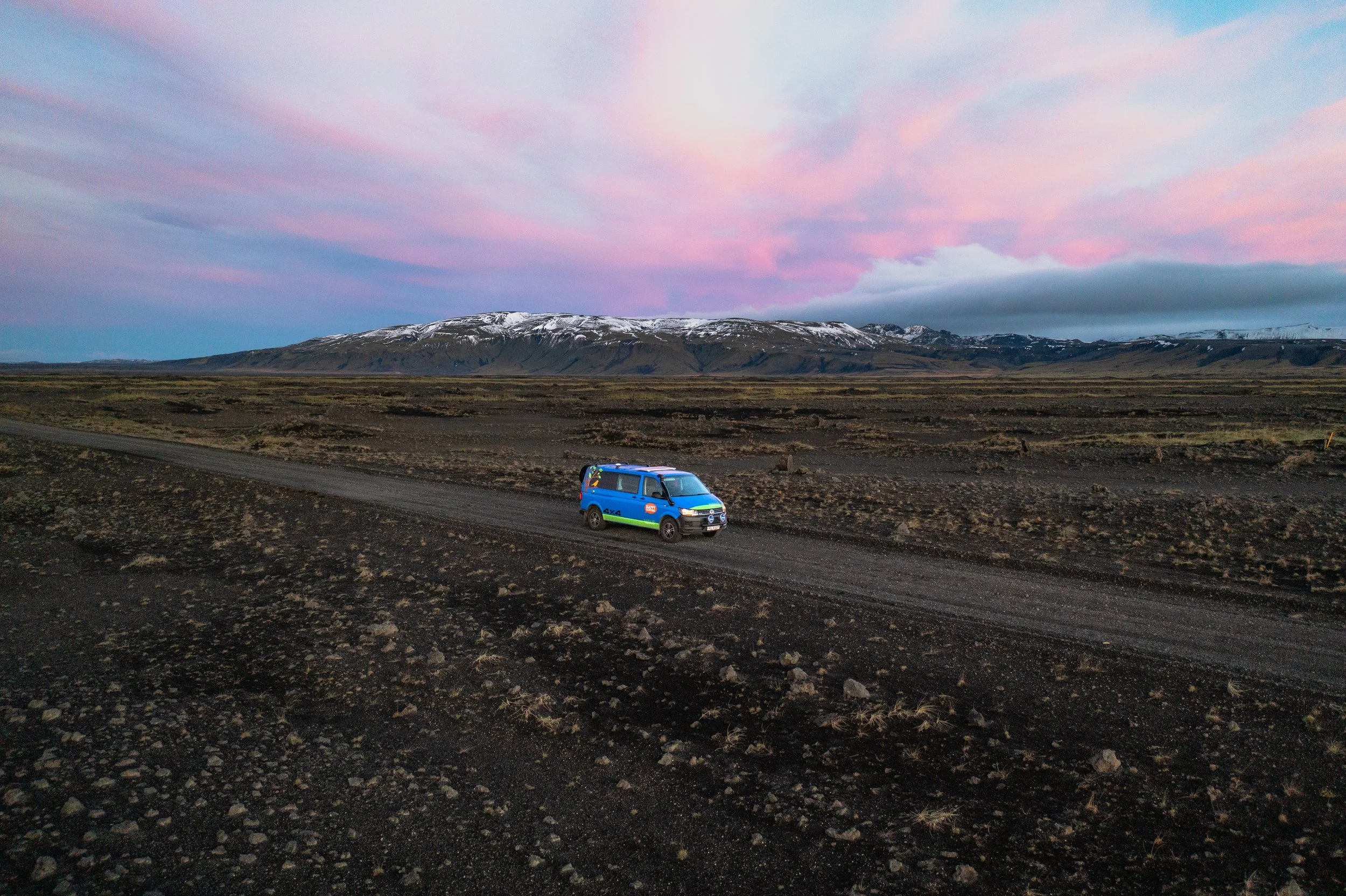 A blue van on a dark dirt road in a barren landscape with mountains in the background and a colorful pink and blue sky above.