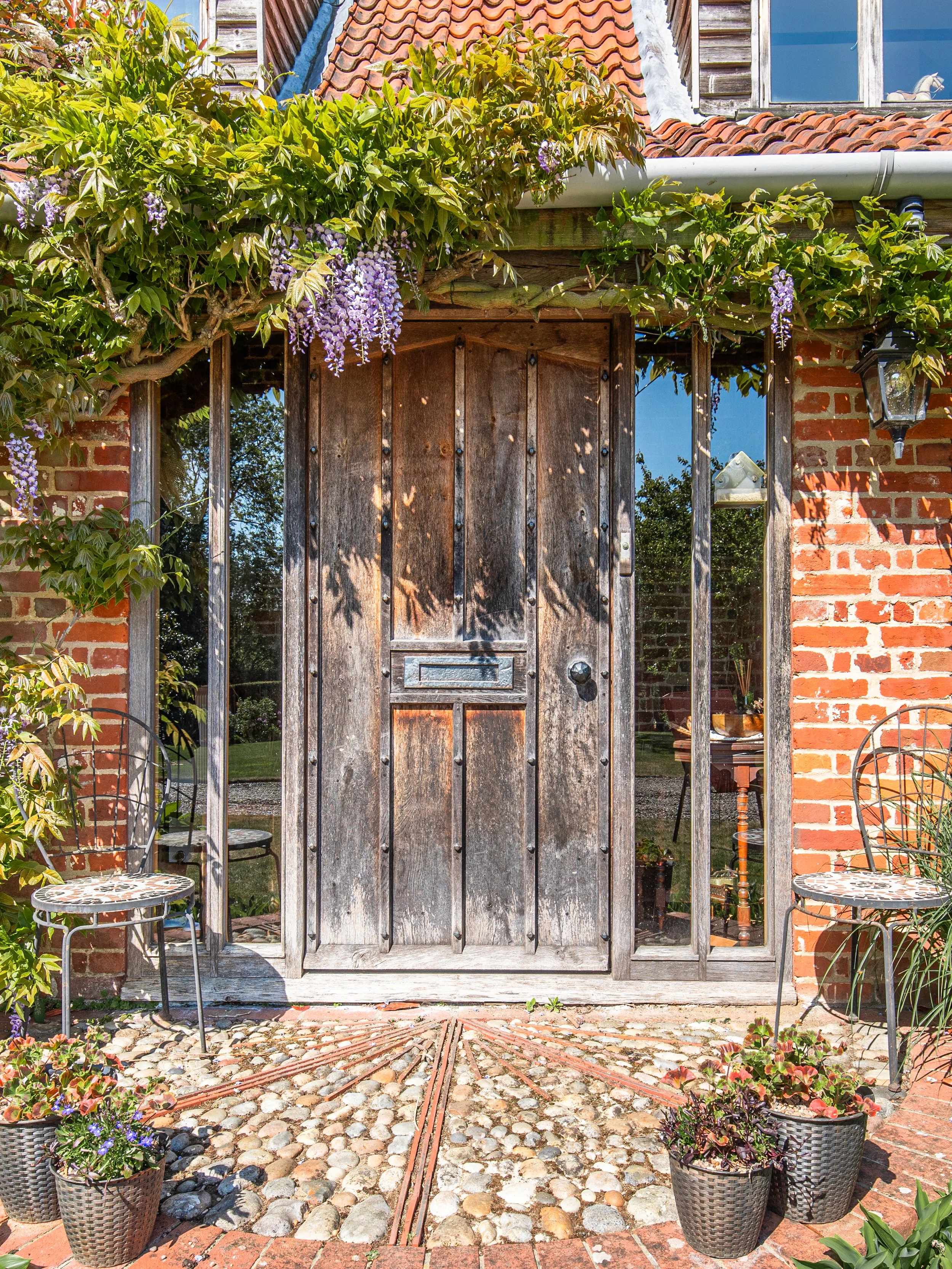 An outdoor scene showcasing a rustic wooden door with a mailbox slot, framed by a metal gate and brick walls. The area is decorated with green plants, flowers, potted plants on either side, two metal chairs, and decorative cobblestone and brick pavem