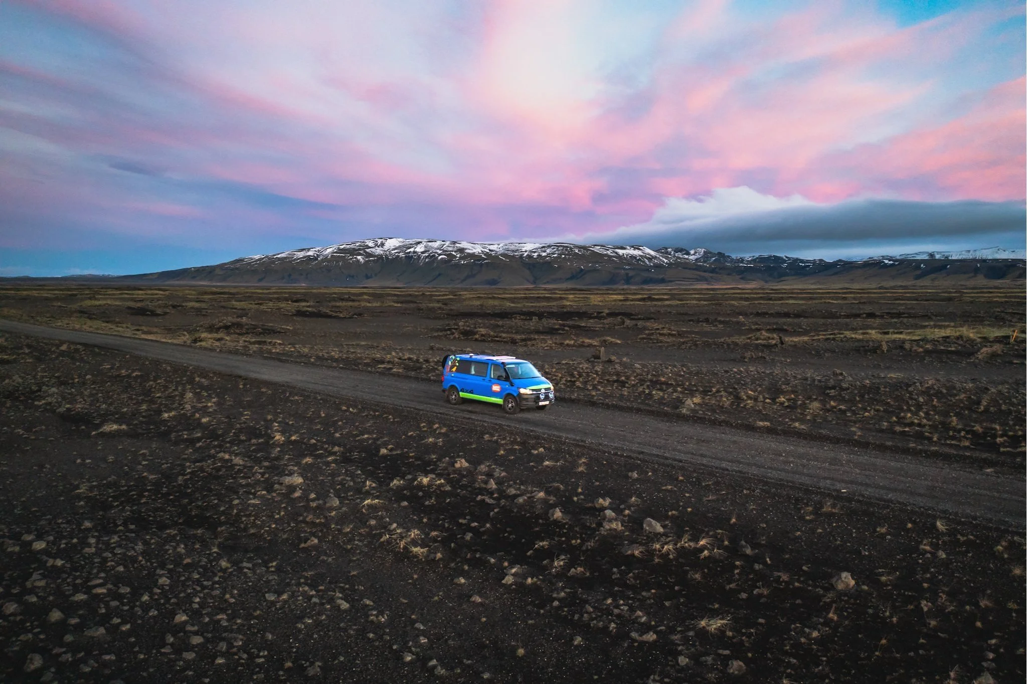 A blue camper van parked on a dirt road in a barren landscape with distant snow-capped mountains and a colorful sunset sky.