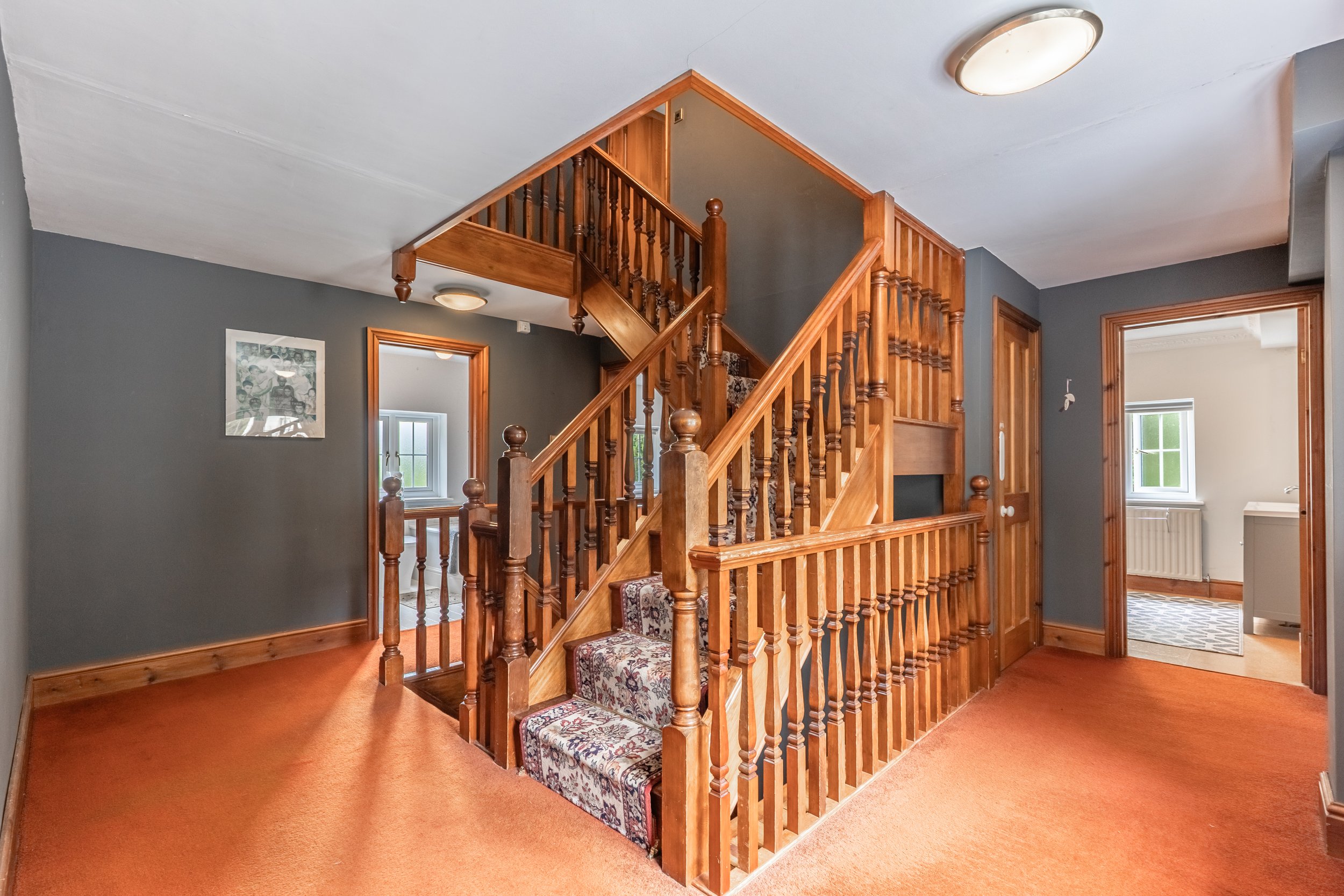 Interior view of a house with a staircase made of wood, leading to the upper floor, with patterned carpet on the stairs, gray walls, and orange carpet flooring.