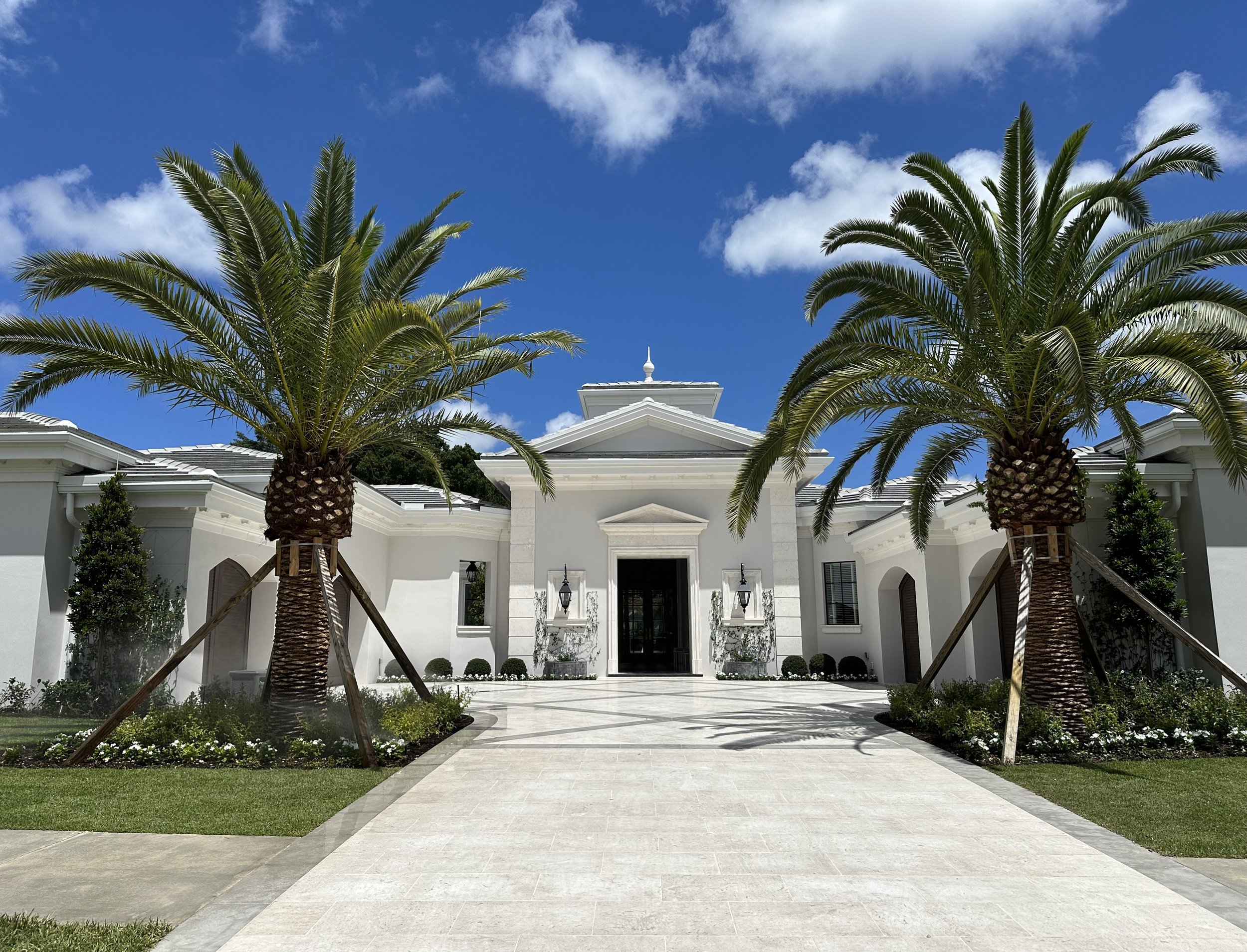 Front view of a luxurious white Caribbean-inspired contemporary architectural designed Florida home with a driveway, two large palm trees, and a bright blue sky with a few clouds.