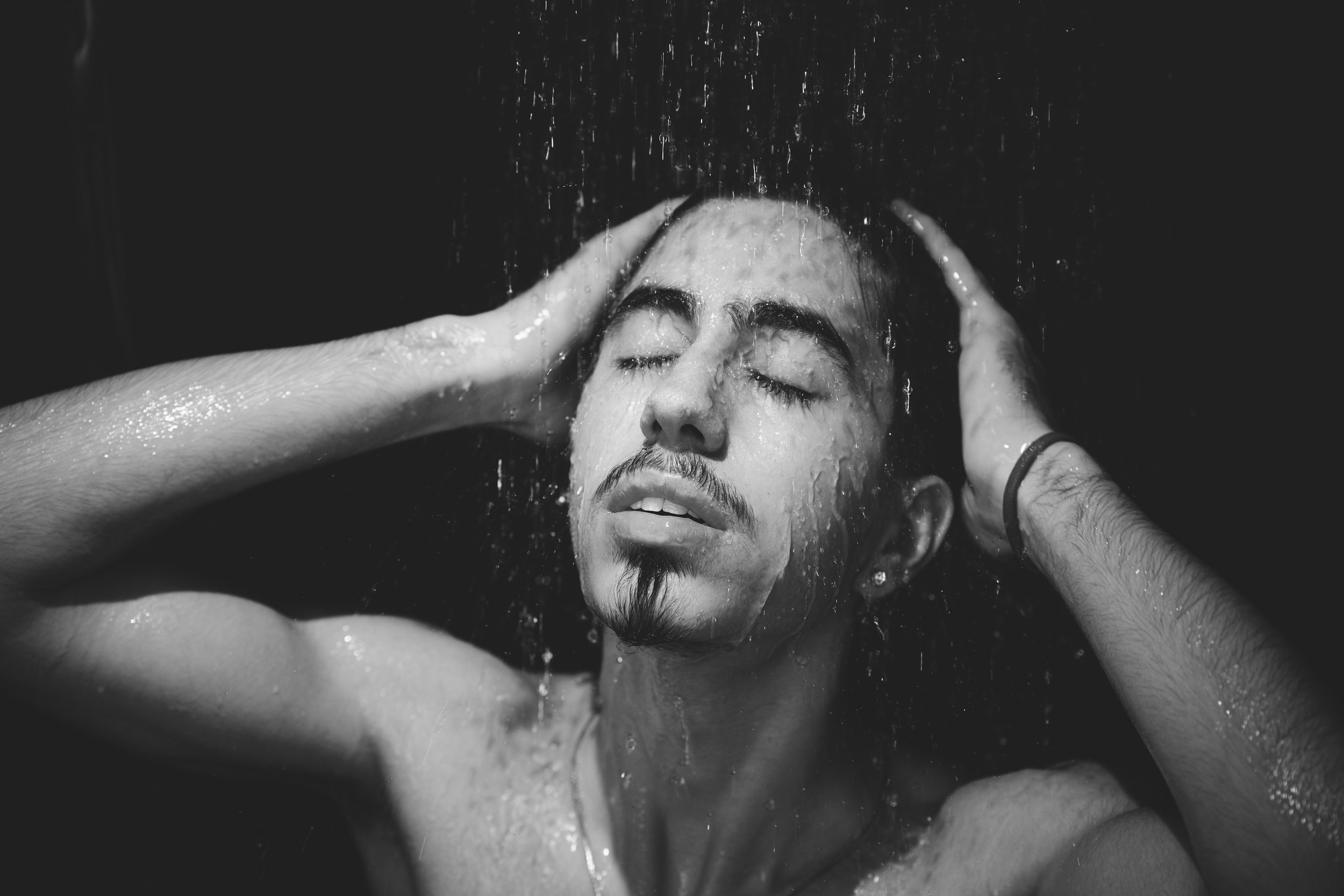 Schwarz-weiß Foto einer Person unter der Dusche, mit geschlossenen Augen und Händen am Kopf.