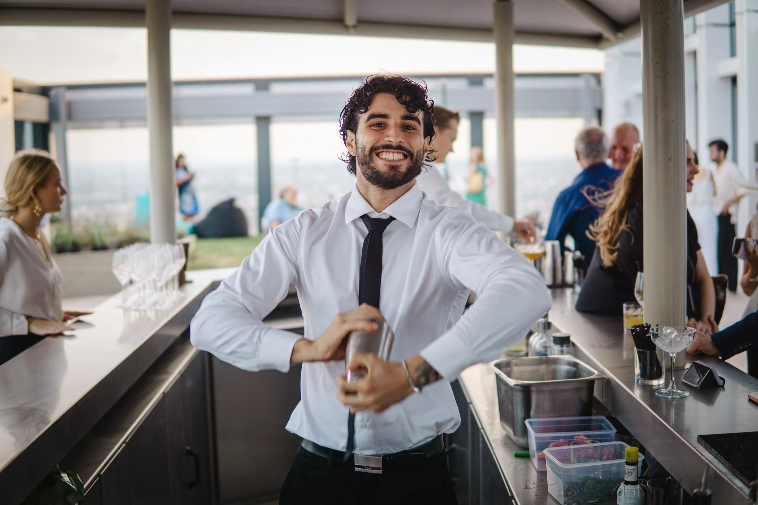 A smiling male bartender with dark curly hair and a beard, wearing a white shirt and black tie, standing behind a bar at an outdoor rooftop venue.