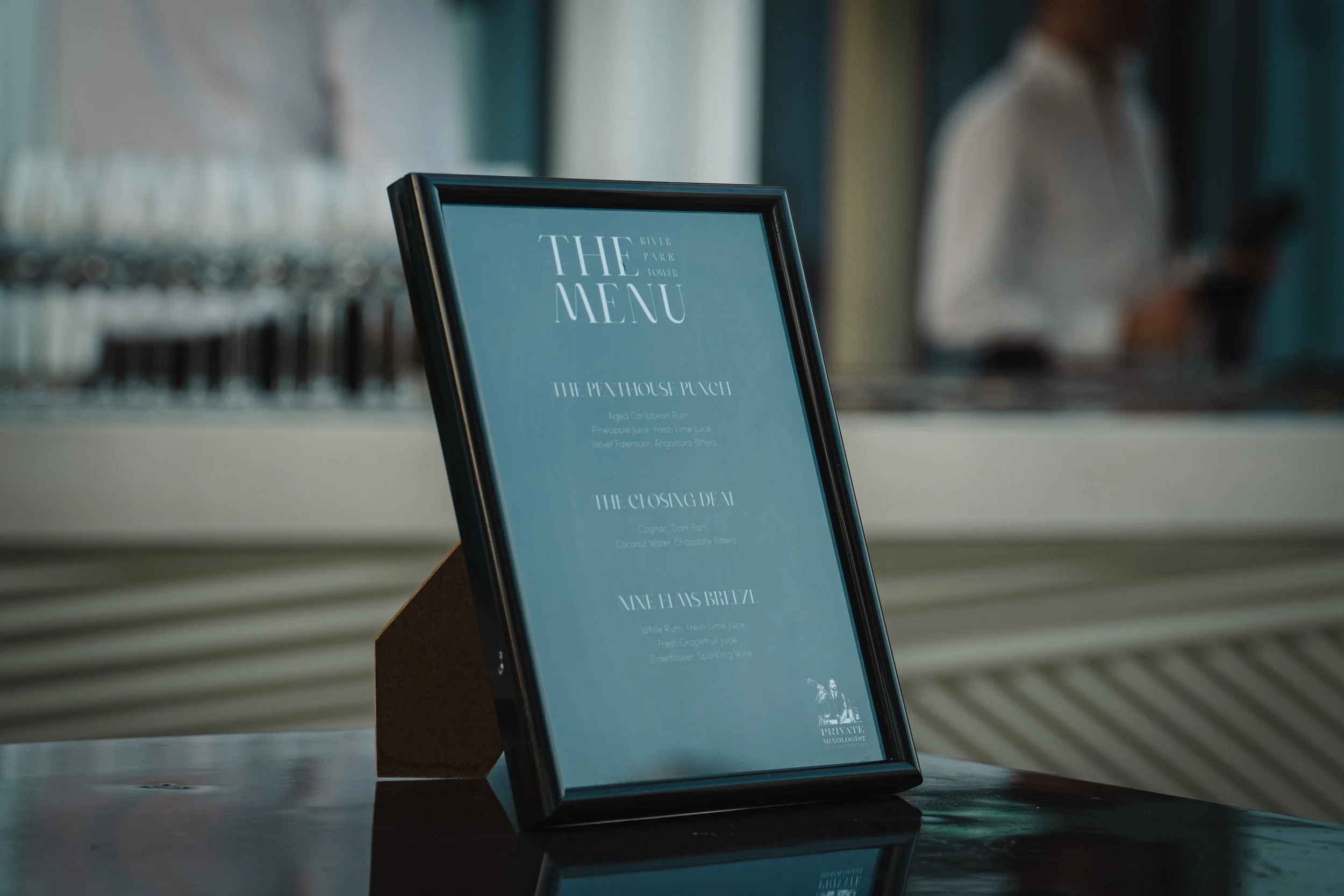 Framed menu displayed on a table at a bar or restaurant, with a person in the background using a cell phone.