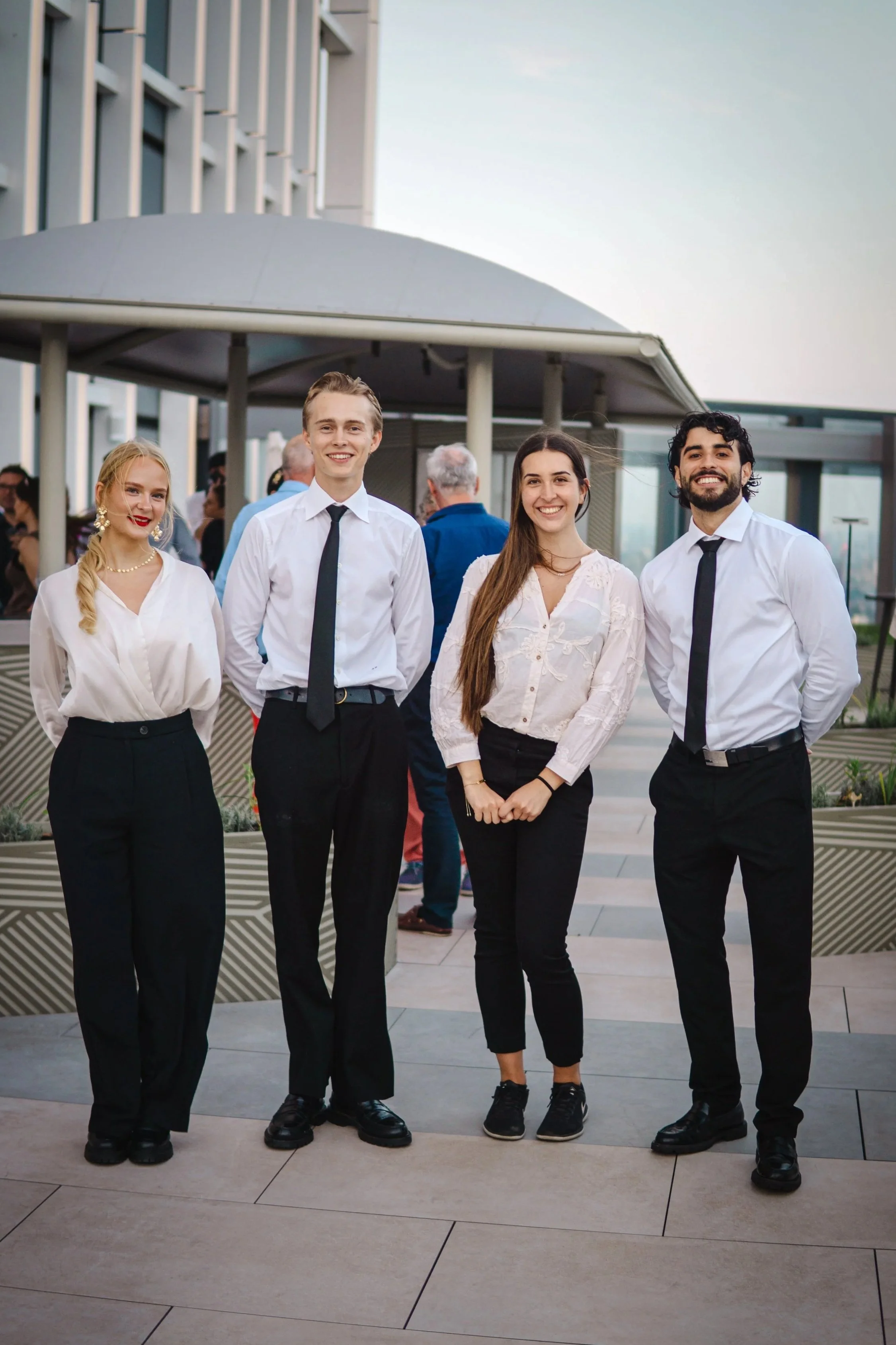 Four young adults in business casual attire posing outdoors at a social event, with a group of people in the background and a modern building nearby.