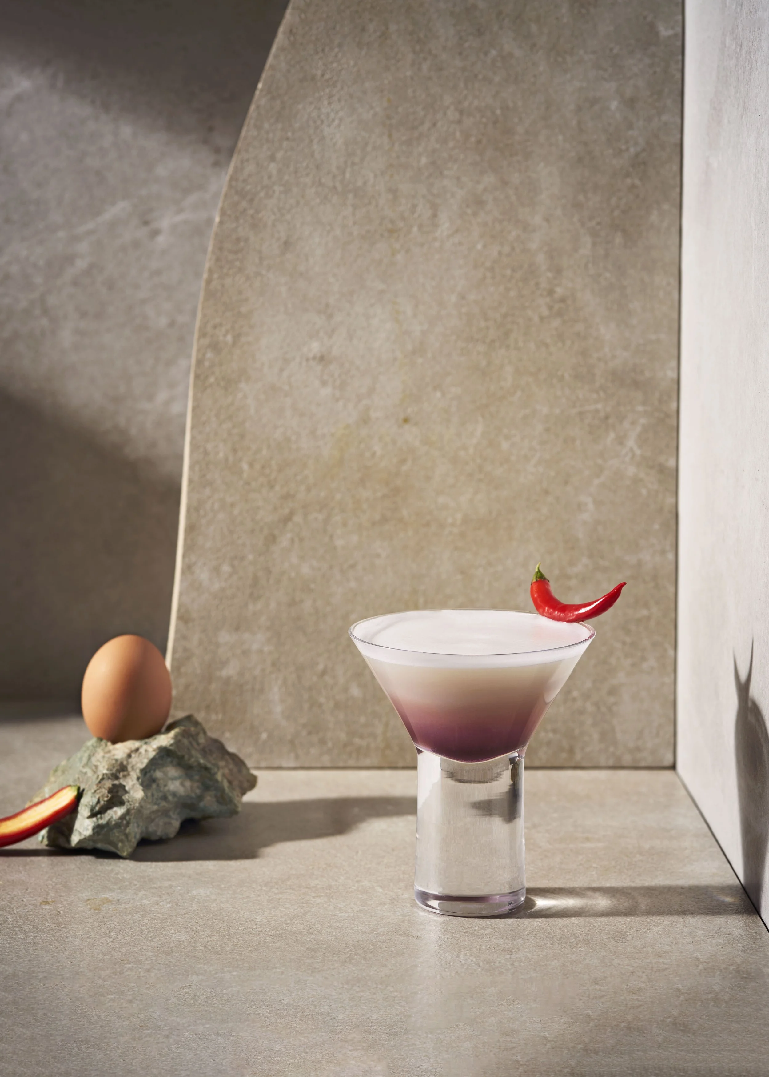 Elegant cocktail with foam topped with a red chili pepper, next to an egg and a rock on a minimalist stone surface.
