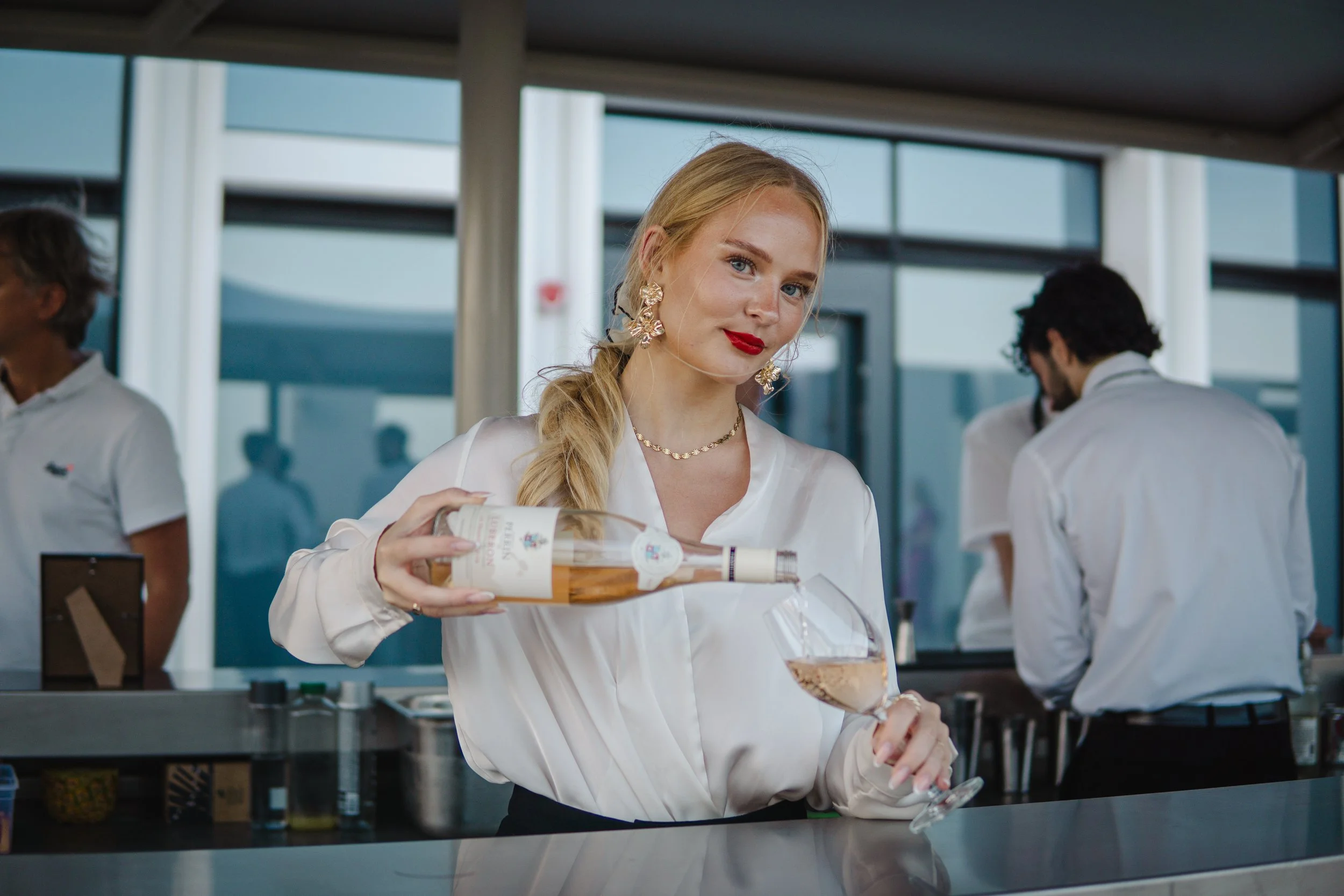 A woman with blonde hair, red lipstick, and gold jewelry pours rosé wine from a bottle into a glass at a bar or restaurant.