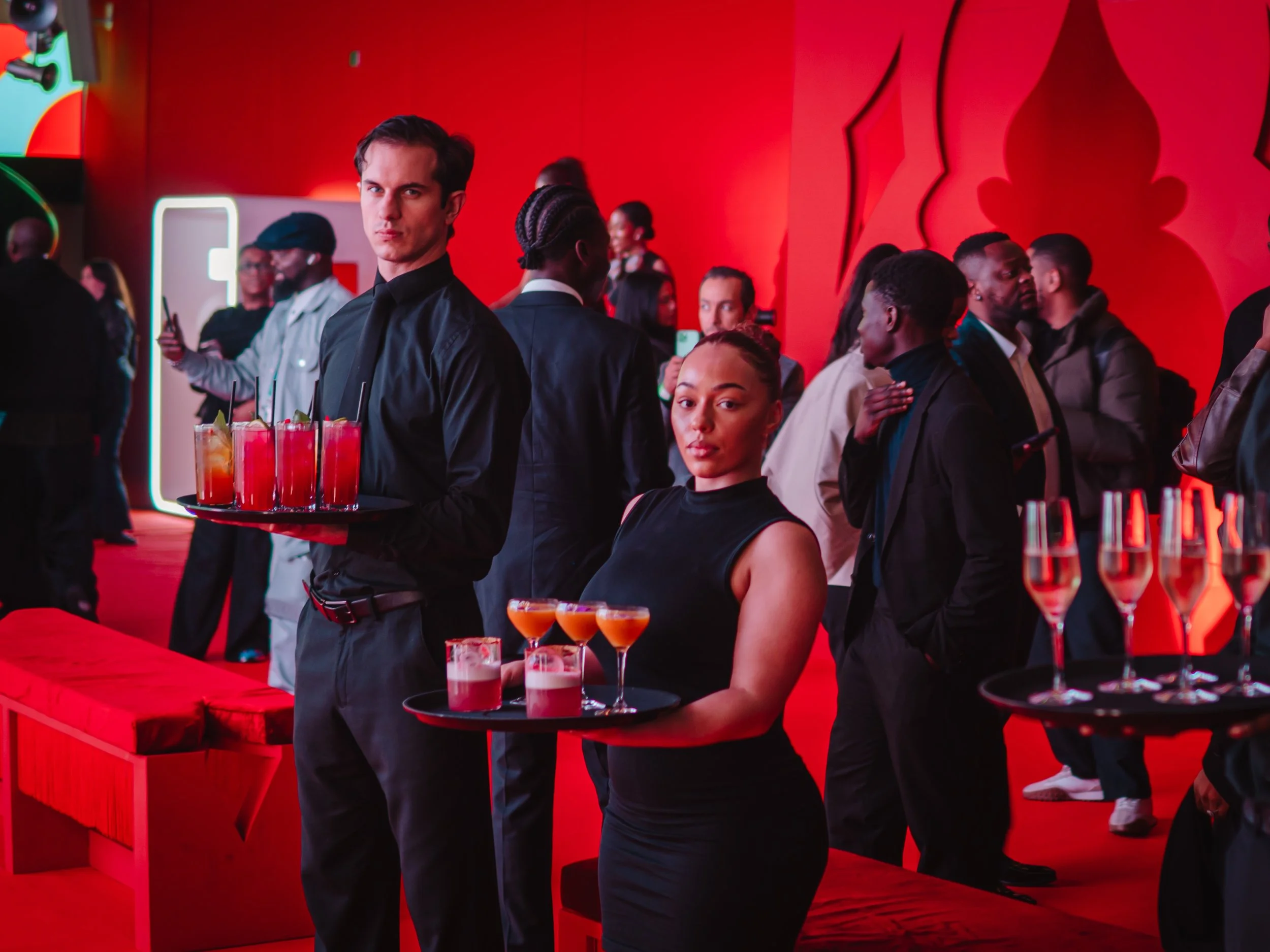 A bartender and a waitress standing behind a red counter with drinks in a bustling, stylish bar or nightclub with red walls and a crowd of people socializing.