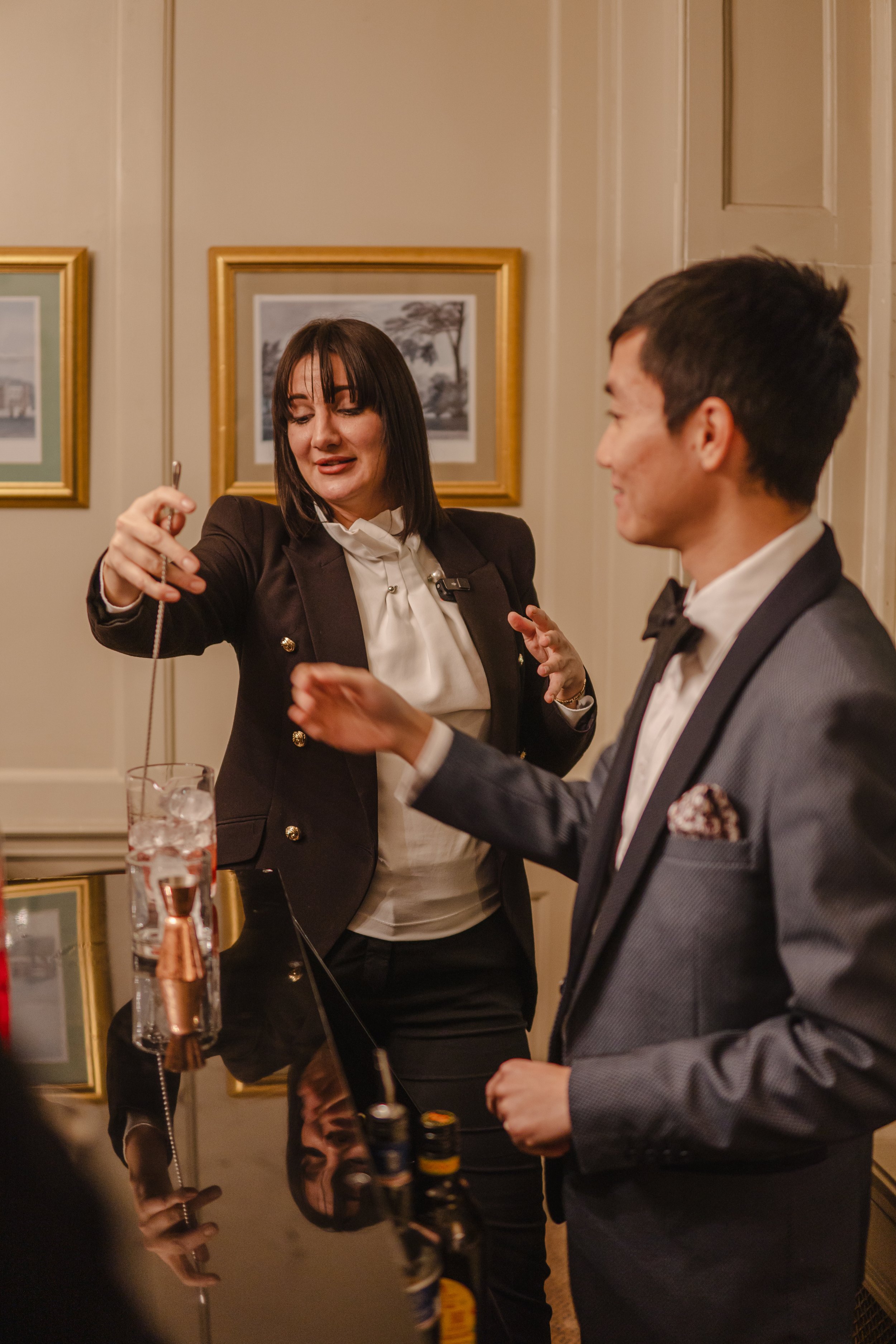 A woman in a black blazer and white blouse pouring a drink into a glass held by a man in a gray tuxedo with a black bow tie, standing at a bar in a room with framed black-and-white pictures on the wall.