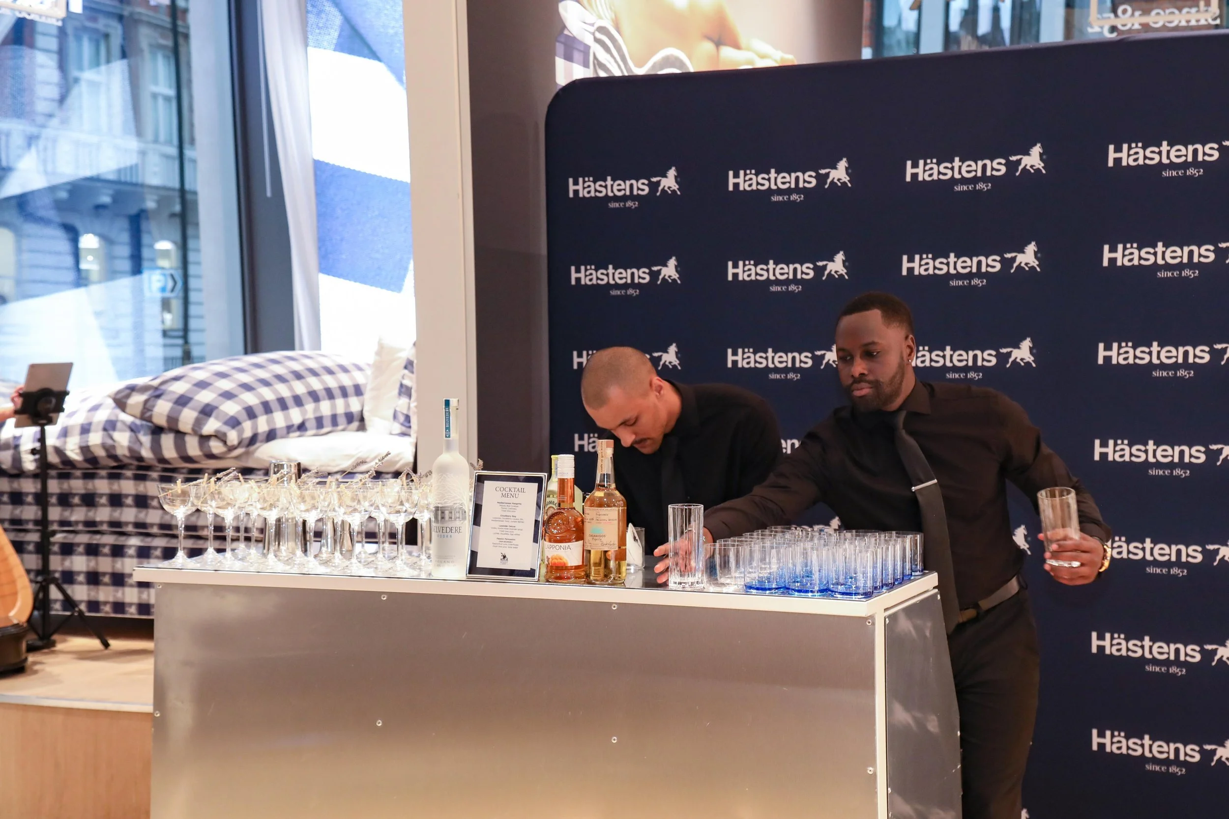 Two bartenders preparing drinks behind a table with glasses and bottles at an event with a Hästens backdrop, a bed with checkered pillows visible in the background.