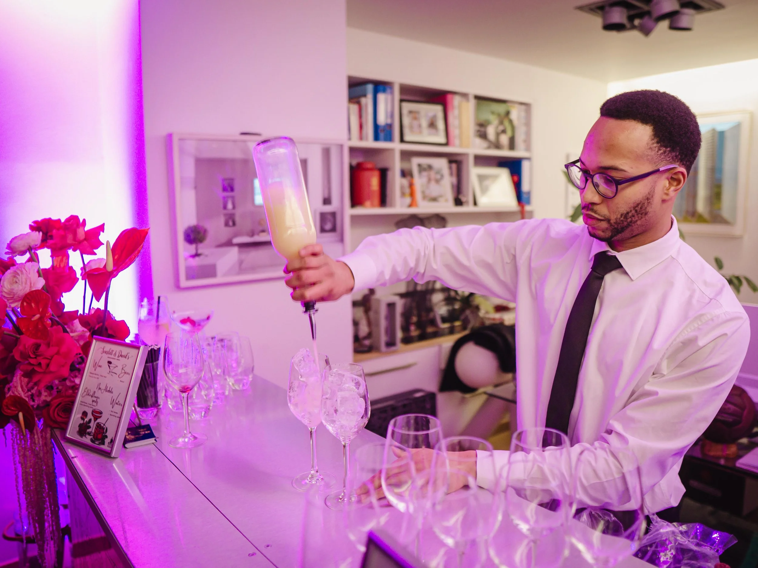 A man in a white shirt, black tie, and glasses is preparing drinks, pouring a creamy liquid from a bottle into a glass with ice, at a table set with various glasses, a bouquet of pink flowers, and a sign. The setting appears to be a decorated indoor space with bookshelves in the background.