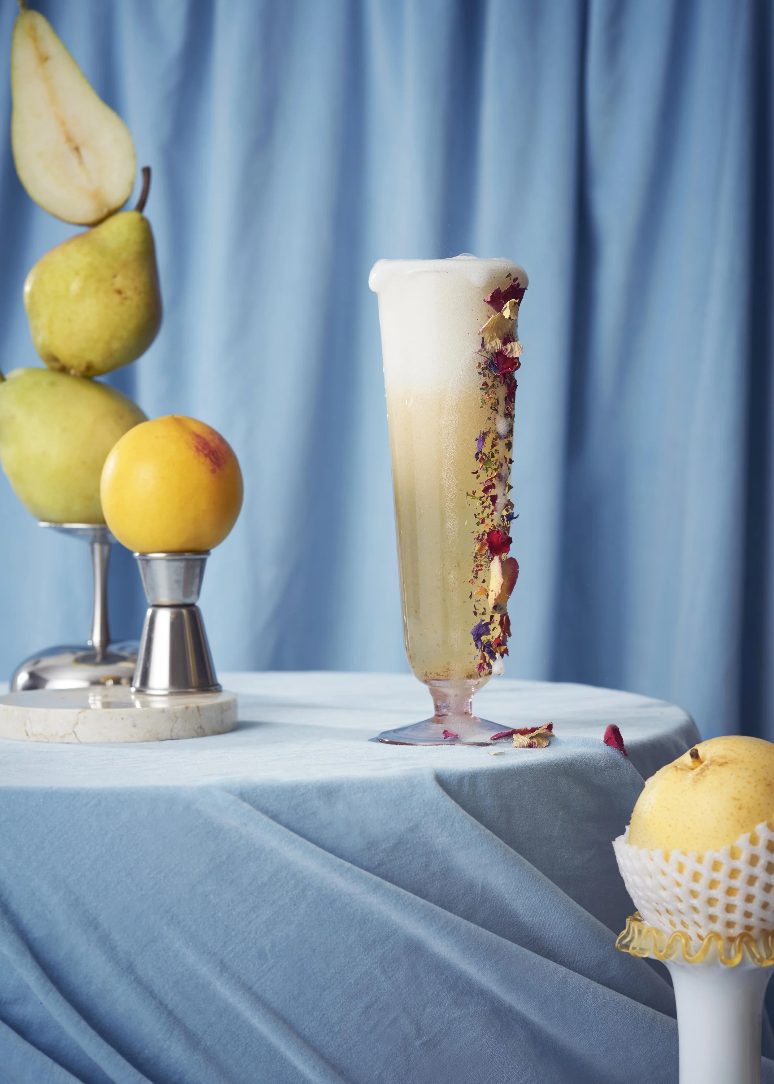 A tall glass of foamy drink with floral decorations sits on a light blue draped tablecloth, alongside a stack of assorted fruits including pears and peach on a metal stand. Background features a light blue curtain.