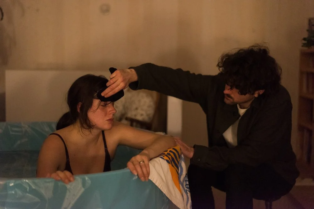 Une femme est en train de recevoir un bain dans une baignoire, un homme la touche doucement sur le front avec une main, comme s'il la soignait ou la réconfortait.