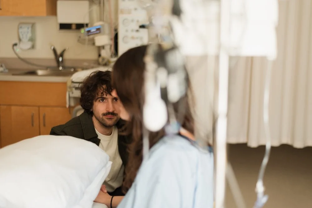 Une femme hospitalisée avec un cathéter, un homme assis à côté dans une chambre d'hôpital.
