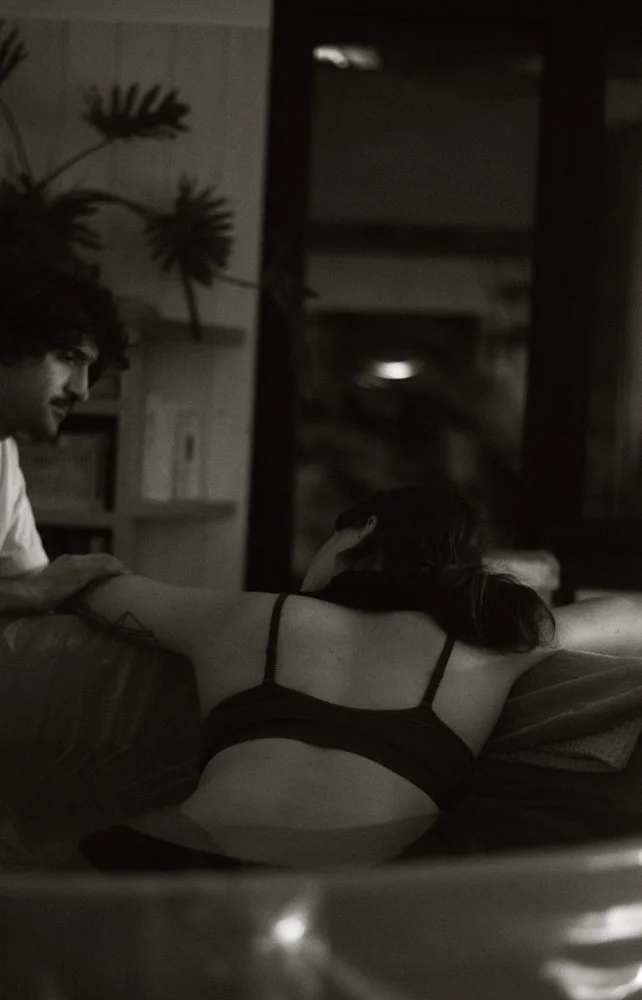 Une scène intime montrant un homme et une femme dans une pièce sombre, avec la femme allongée sur un lit ou un canapé, vue de dos, portant un vêtement noir, et l'homme assis à ses côtés. La pièce est décorée avec des plantes et des étagères en arrièr