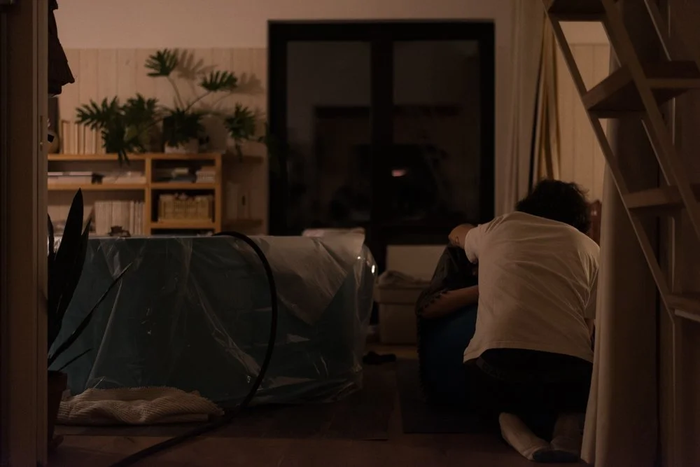 Deux personnes embrassent dans un salon faiblement éclairé avec des meubles en bois, des plantes et une grande fenêtre ou porte-fenêtre.