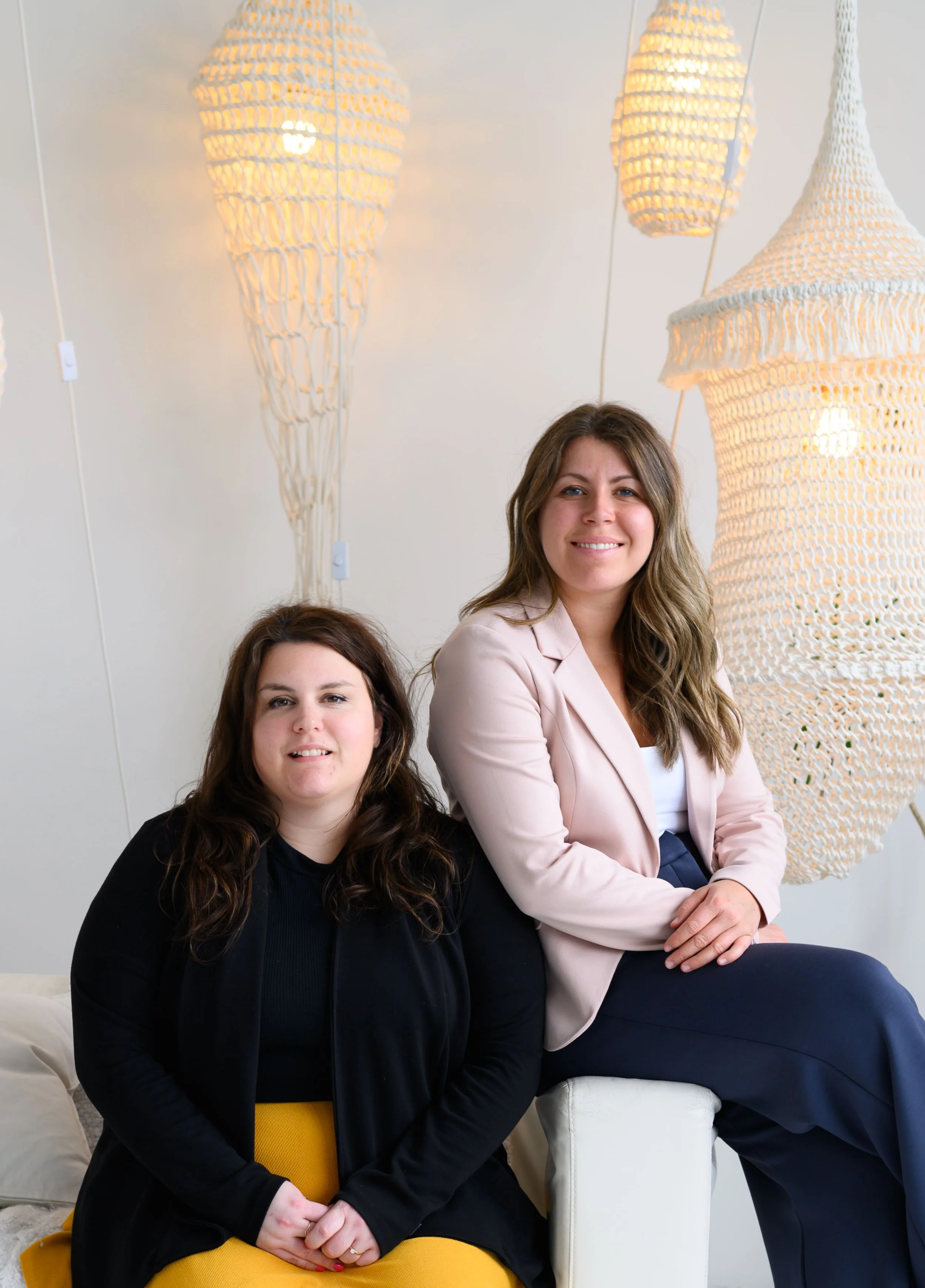 Deux femmes posent dans un intérieur lumineux avec des suspensions en rotin suspendues au plafond.