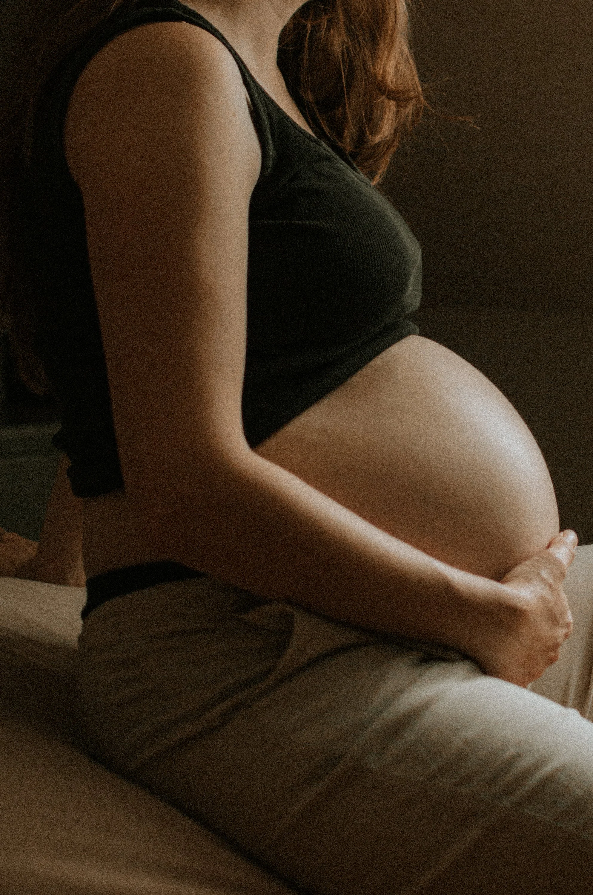 Une femme enceinte assise sur un lit, portant un haut noir, tenant son ventre avec une main.