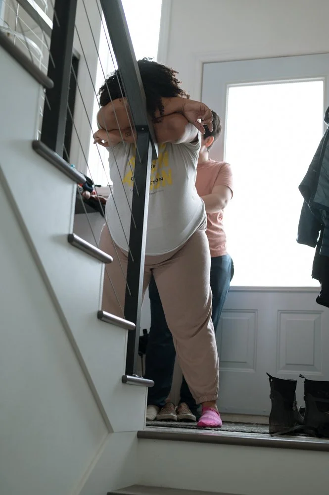 Personne qui descend les escaliers, en arrière-plan un autre individu et un mur avec une grande fenêtre, vêtements décontractés.